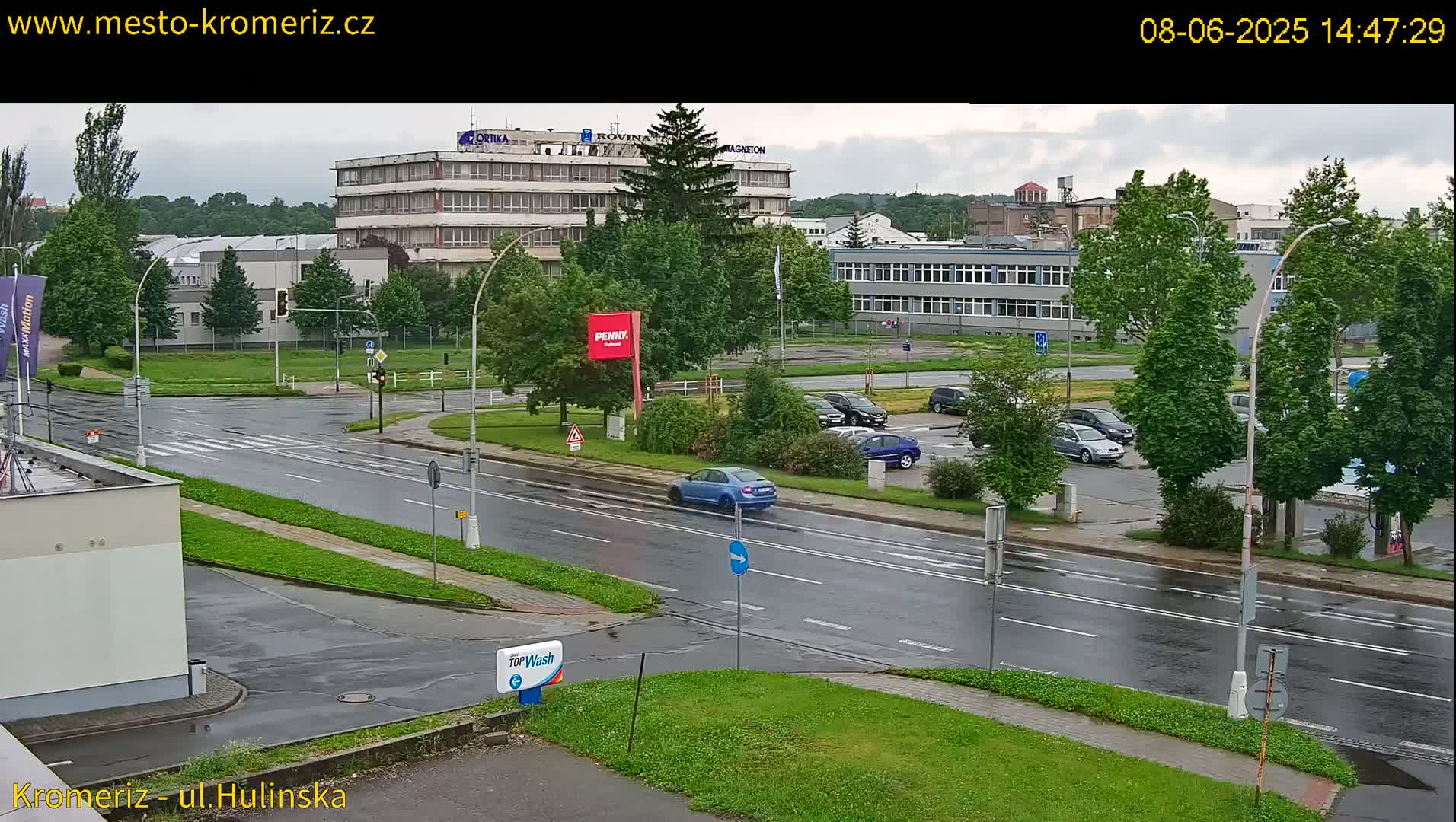 A wet, overcast street scene shows several cars parked in a lot next to a multi-story building, with a few cars driving on the rain-slicked road.