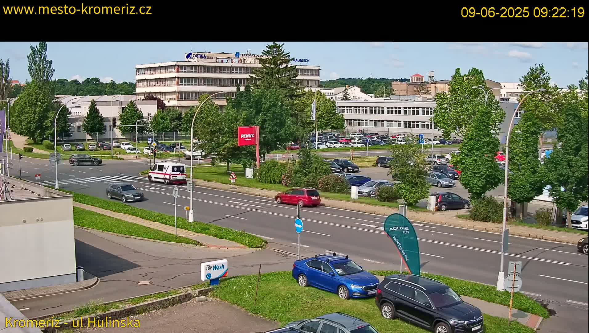 A sunny day reveals a street scene with several cars driving and parked near a large building complex and a car wash.