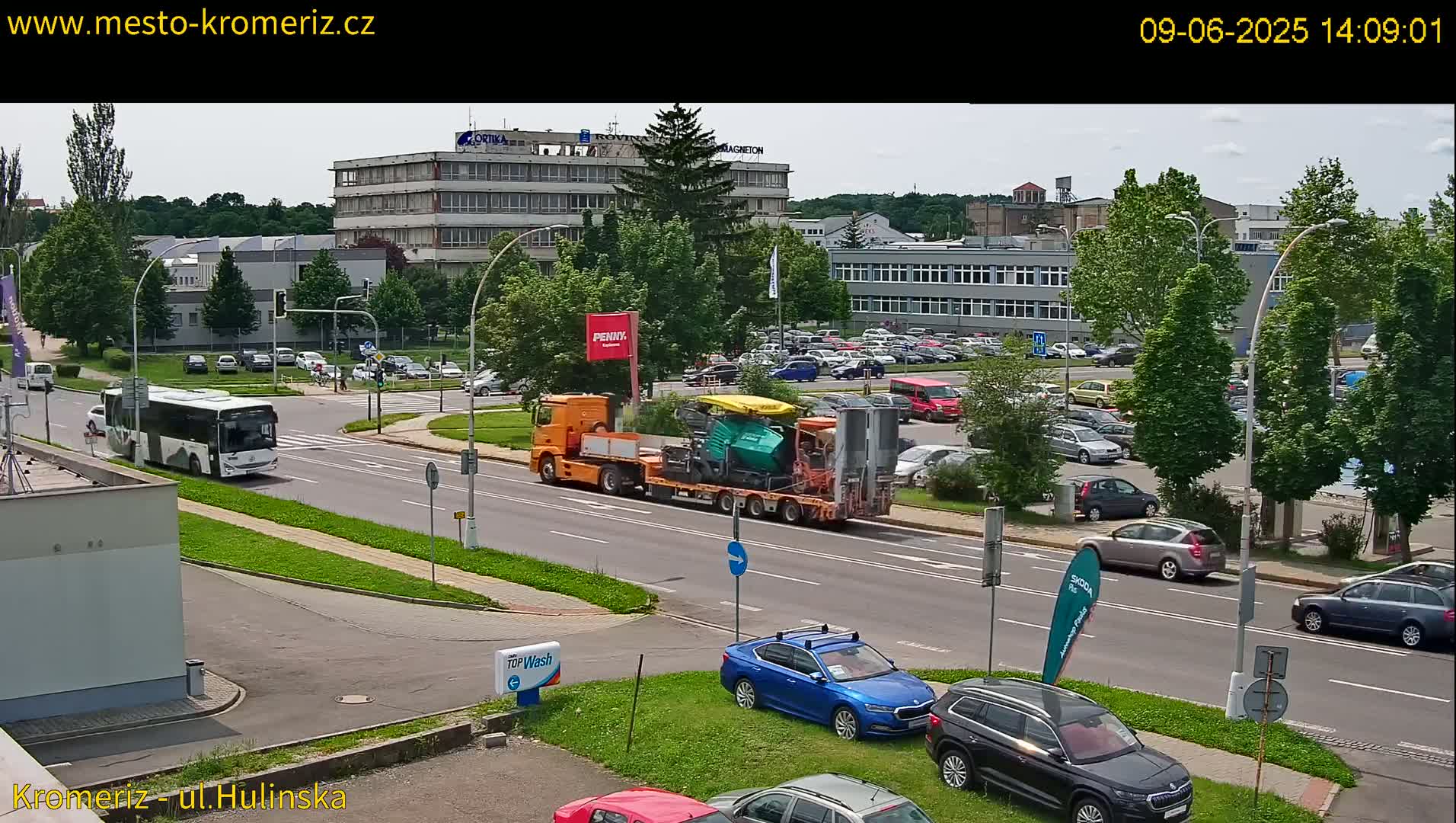 A sunny day shows a flatbed truck carrying large pieces of construction equipment driving on a city street past a large parking lot and several buildings.