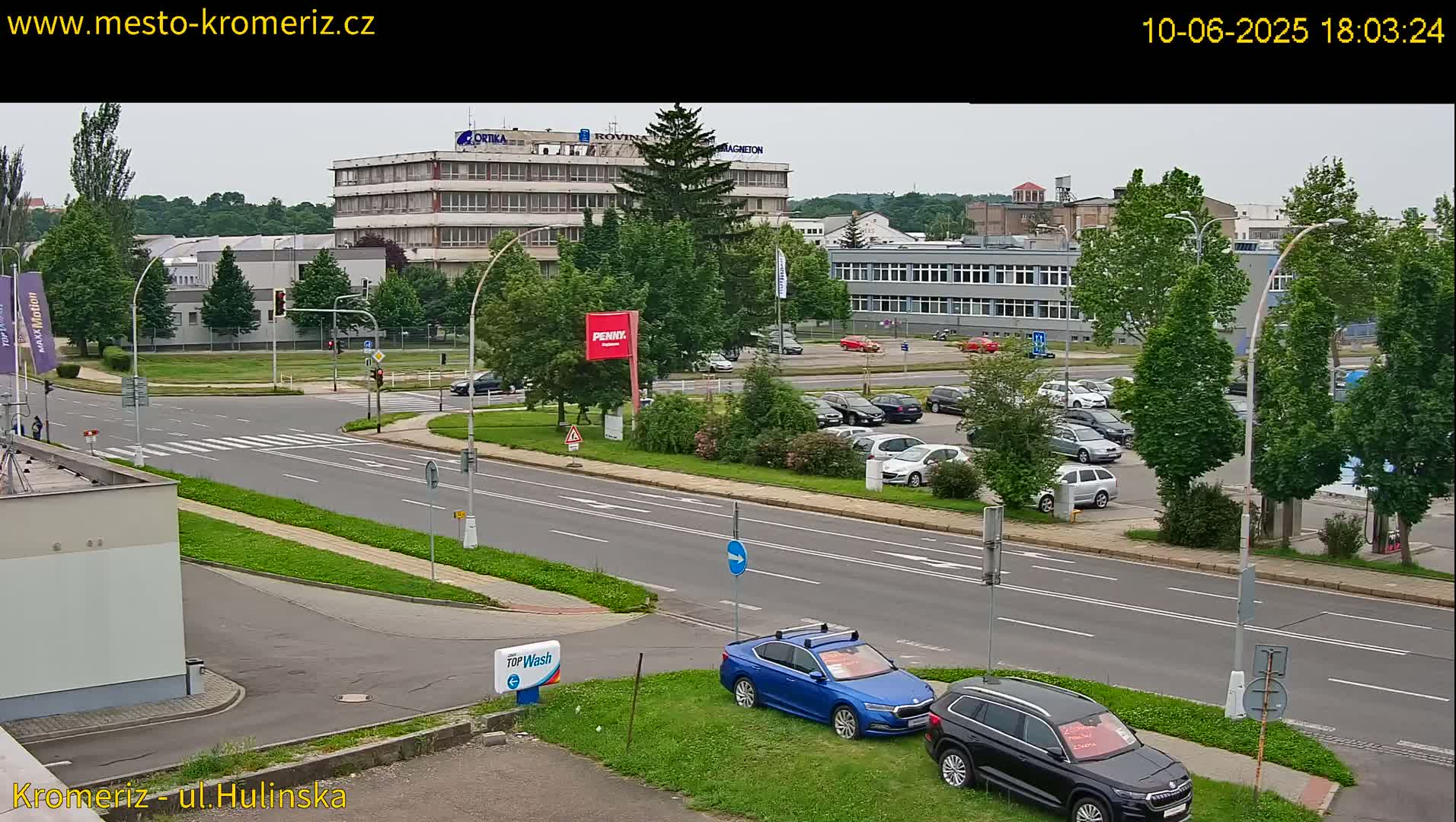 A high-angle, long shot of a city street intersection with several cars parked in a lot and a few cars driving on the road, under an overcast sky.