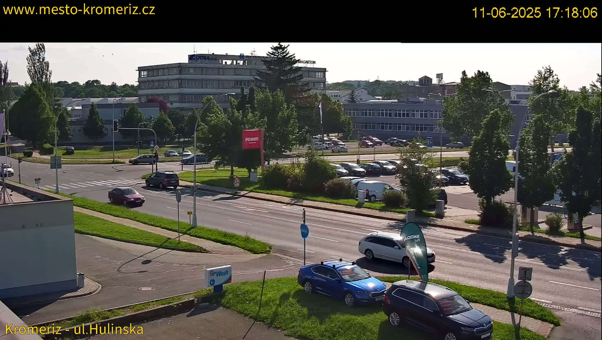 A sunny day shows a multi-lane road with cars driving past several buildings and a parking lot filled with vehicles;  trees and grassy areas surround the road.