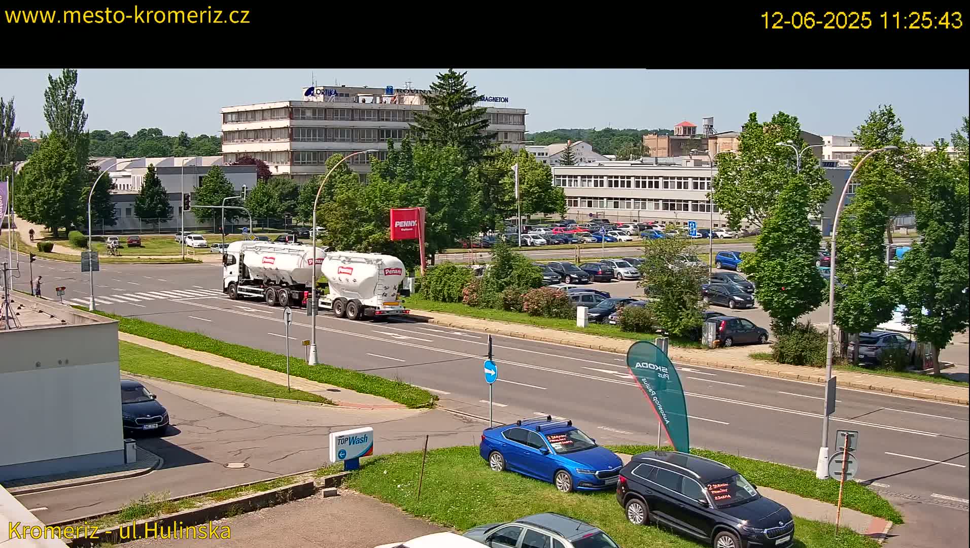 A sunny day shows a large industrial area with a parking lot full of cars, a cement mixer truck driving on a road, and several other vehicles parked nearby.
