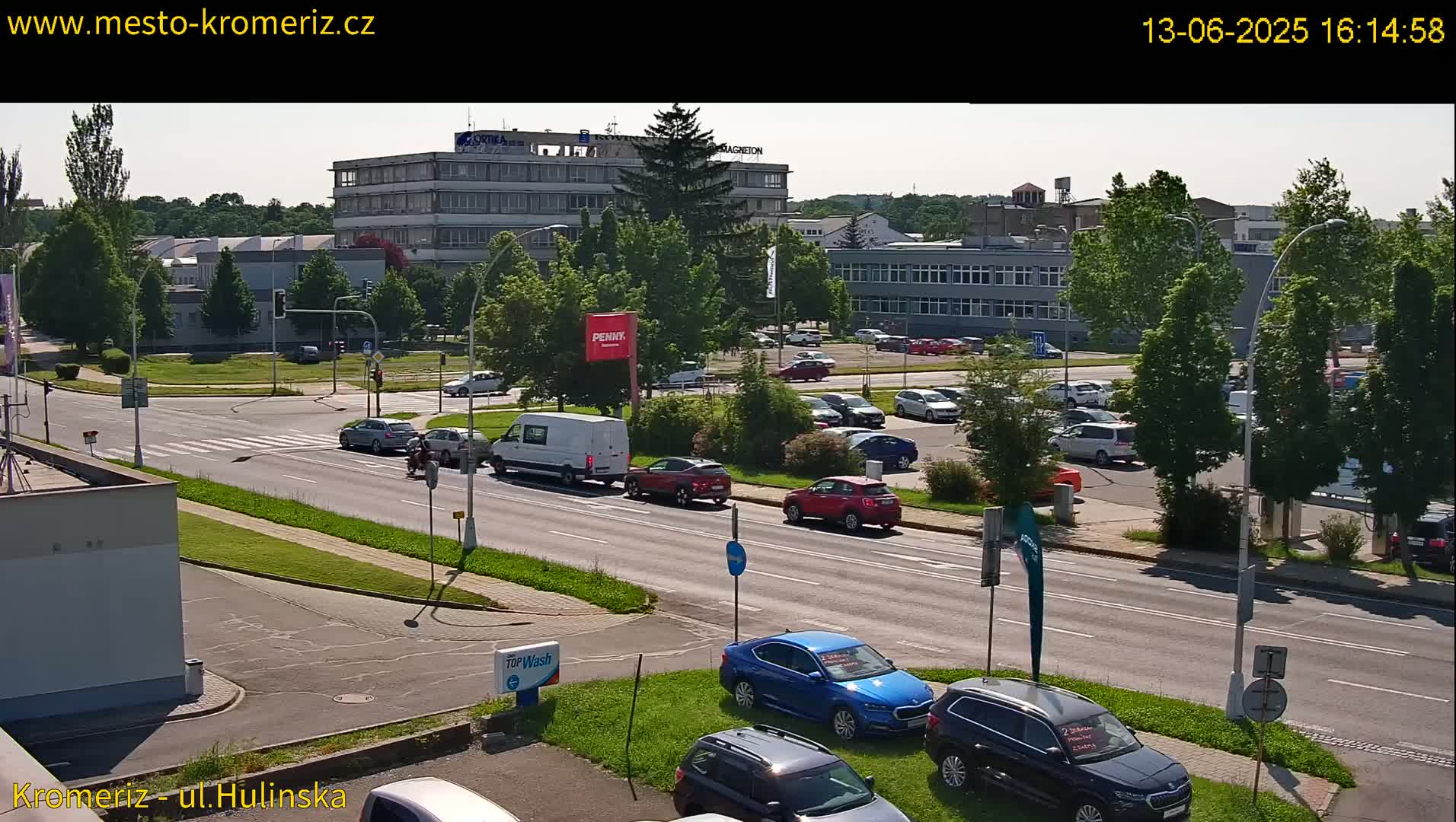 A sunny day reveals a city street scene with several cars parked in a lot and others driving on a multi-lane road, alongside buildings and lush green trees.