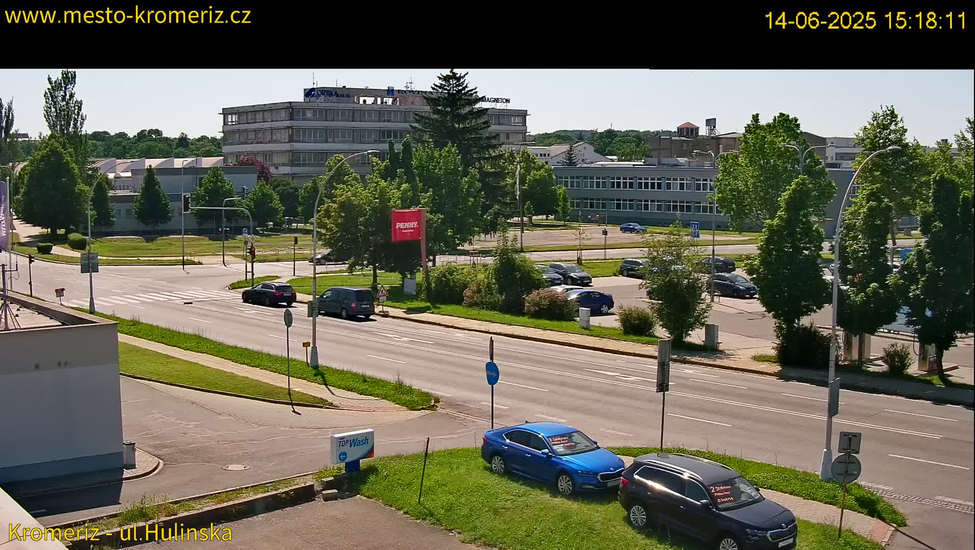 A sunny day reveals a wide road with cars parked along its sides and several buildings in the background, including a multi-story structure.