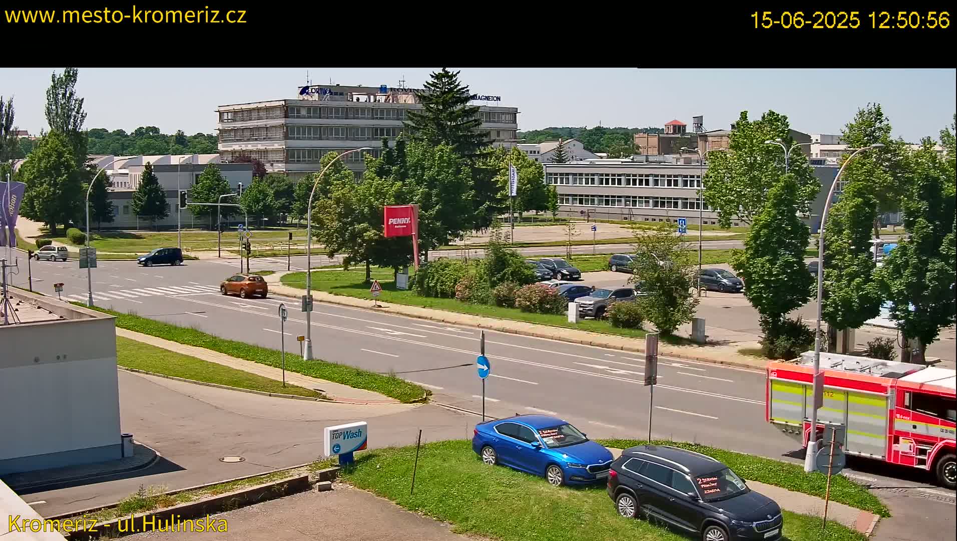 A sunny day shows a street scene with cars parked in a lot and driving on the road, next to buildings and green trees, with a firetruck visible on the right.