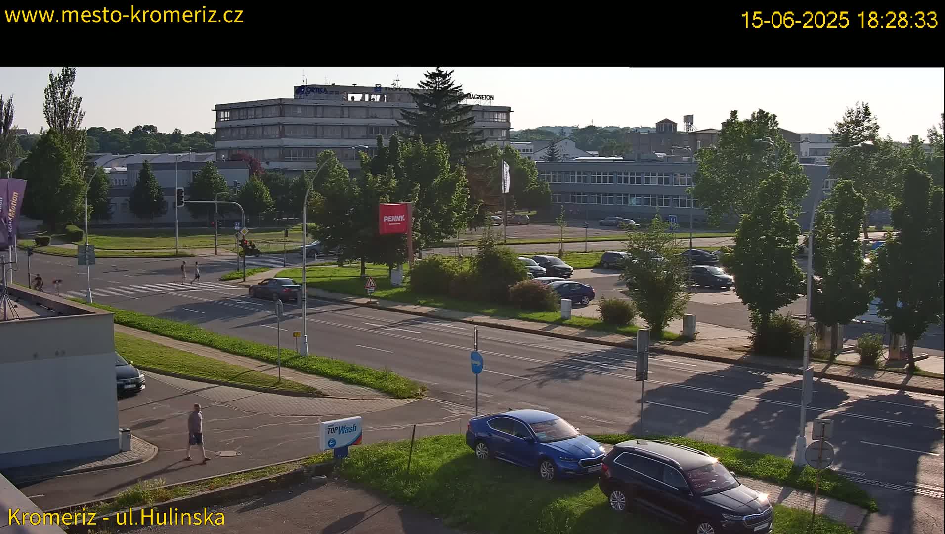 A sunny, late afternoon view of a city street intersection with several cars parked and driving, pedestrians crossing, and buildings in the background.