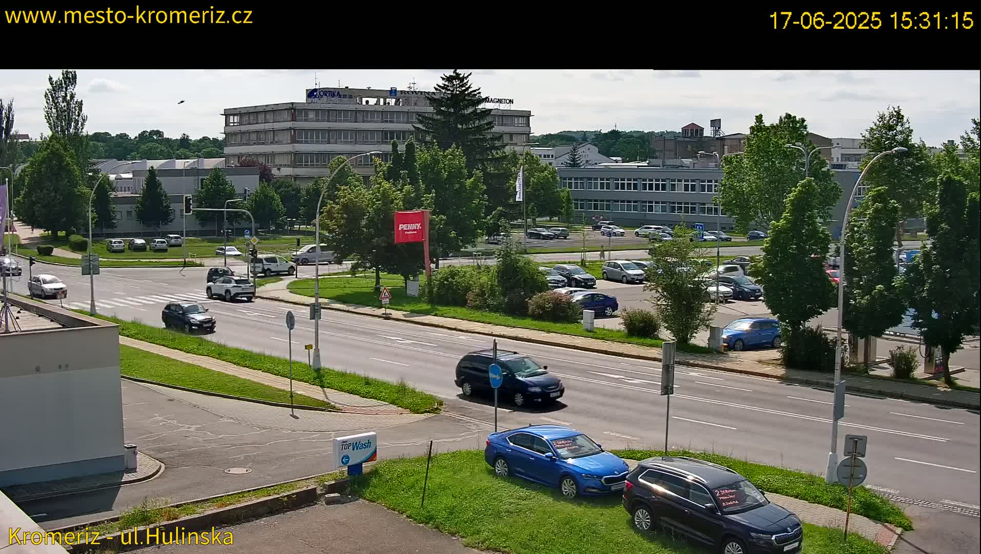 A sunny day reveals a street scene with several cars parked in a lot and driving on a multi-lane road, near several buildings and green spaces.