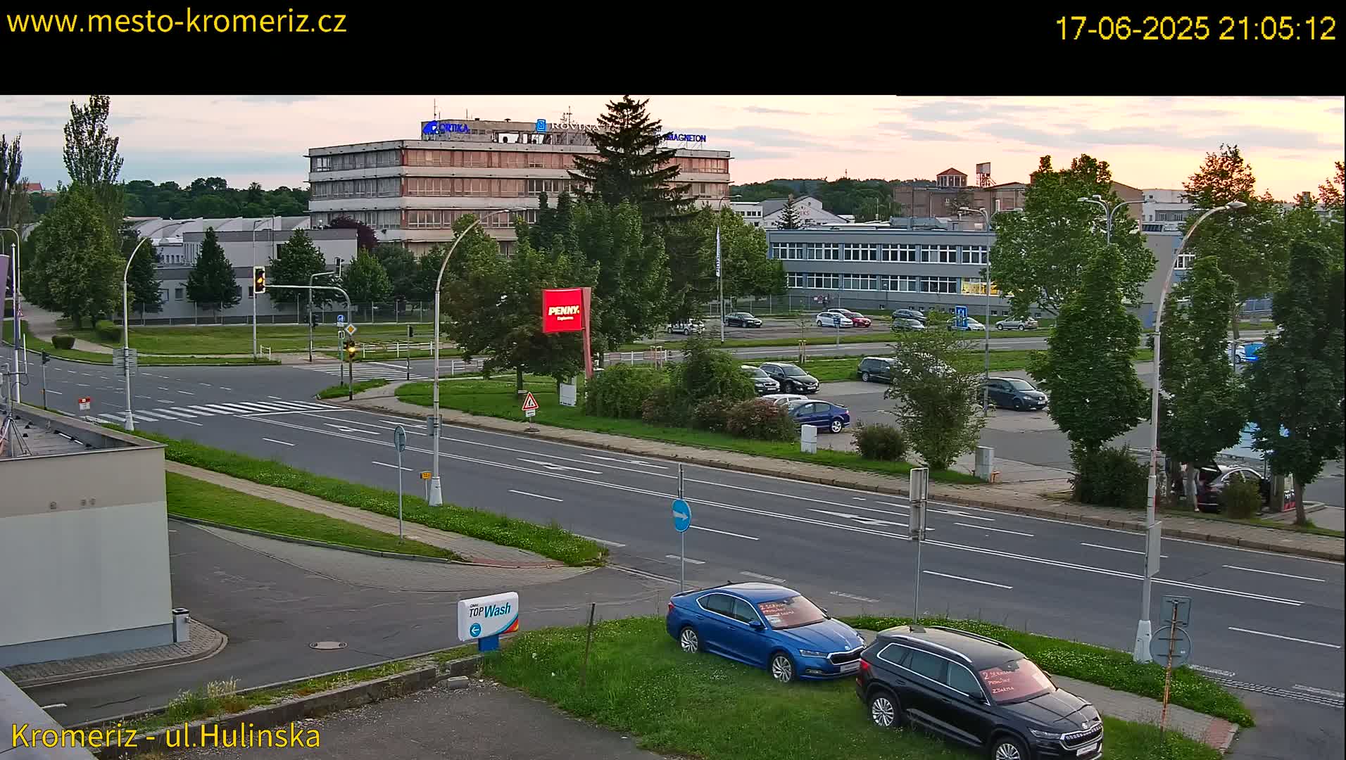 A twilight view of a multi-lane road and parking lot near several buildings, with cars parked and driving, under a partly cloudy sky.