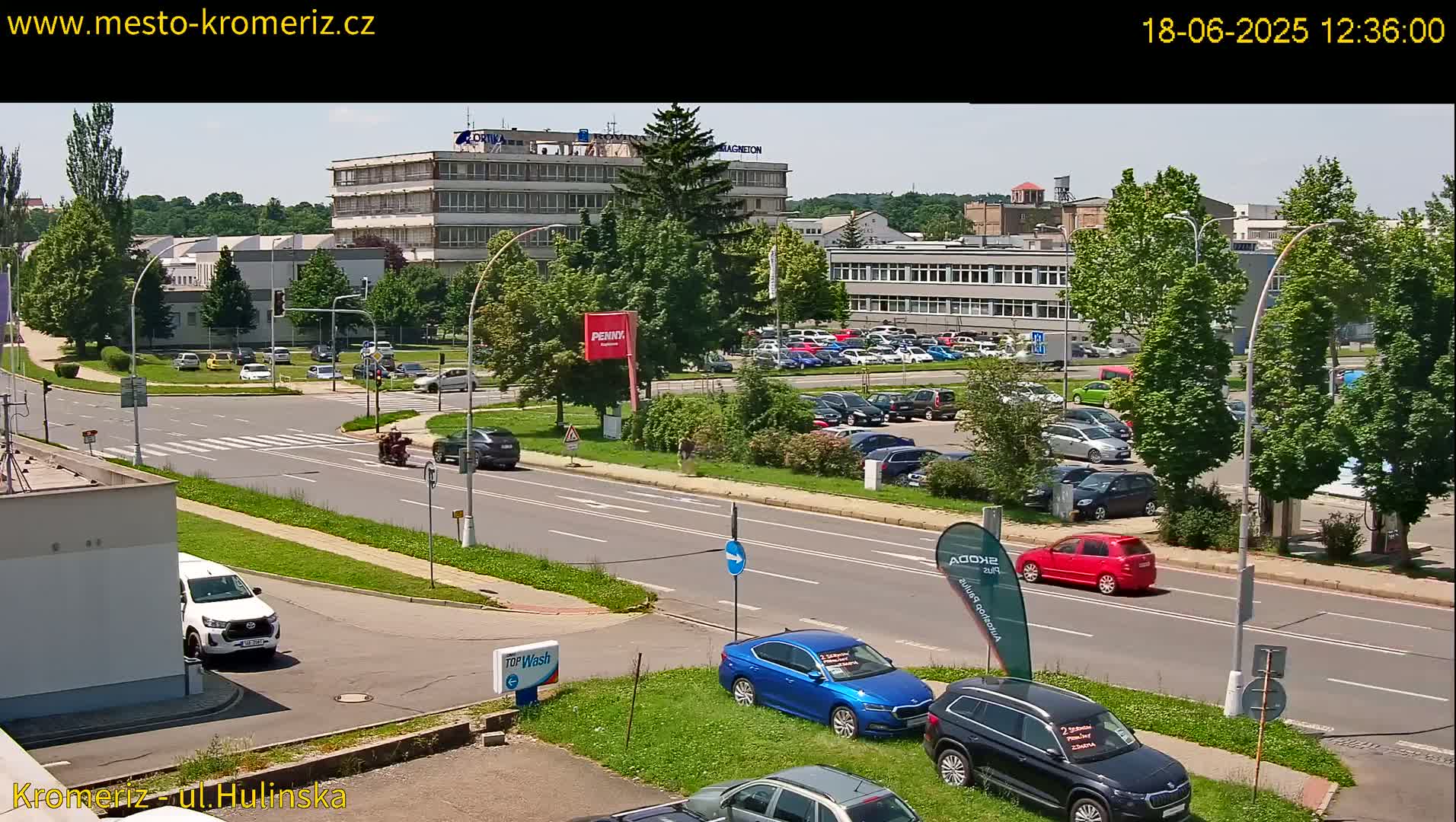 A sunny day view of a street scene with several cars parked in a lot and driving on the road, in front of several multi-story buildings and lush green trees.