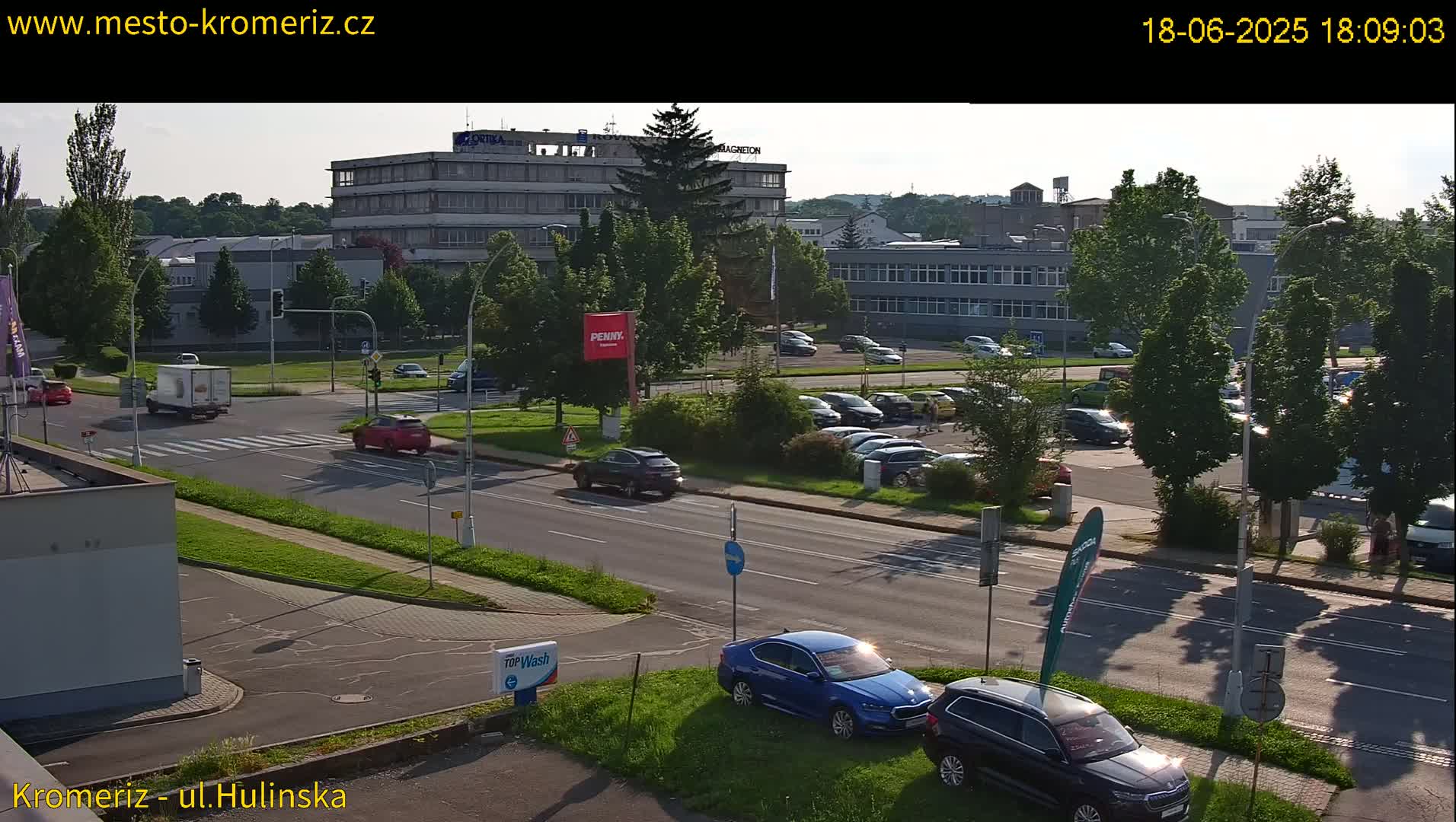 A sunny afternoon scene shows a multi-lane road with cars and a few pedestrians, next to a parking lot filled with vehicles, and in the background, several industrial-looking buildings surrounded by green trees.
