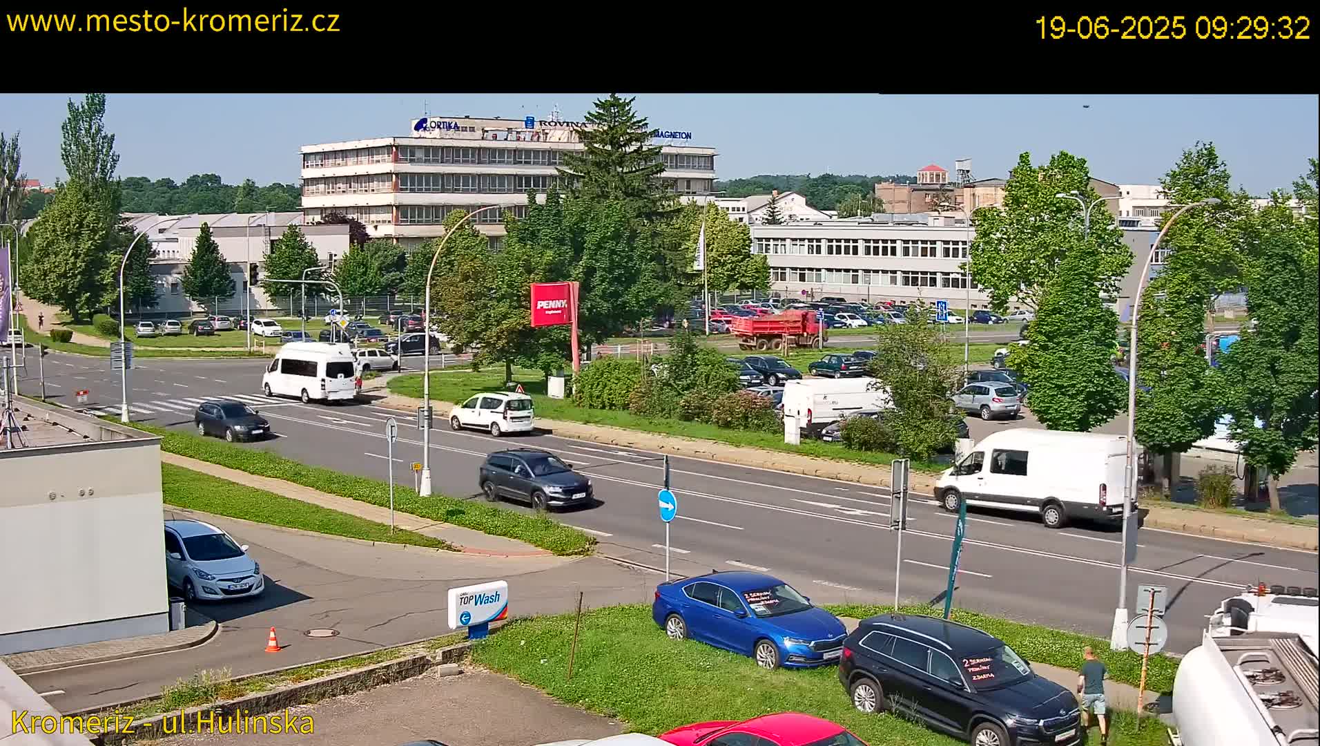 A sunny day shows a multi-lane road with several cars and vans driving past buildings, and parked cars in a lot.