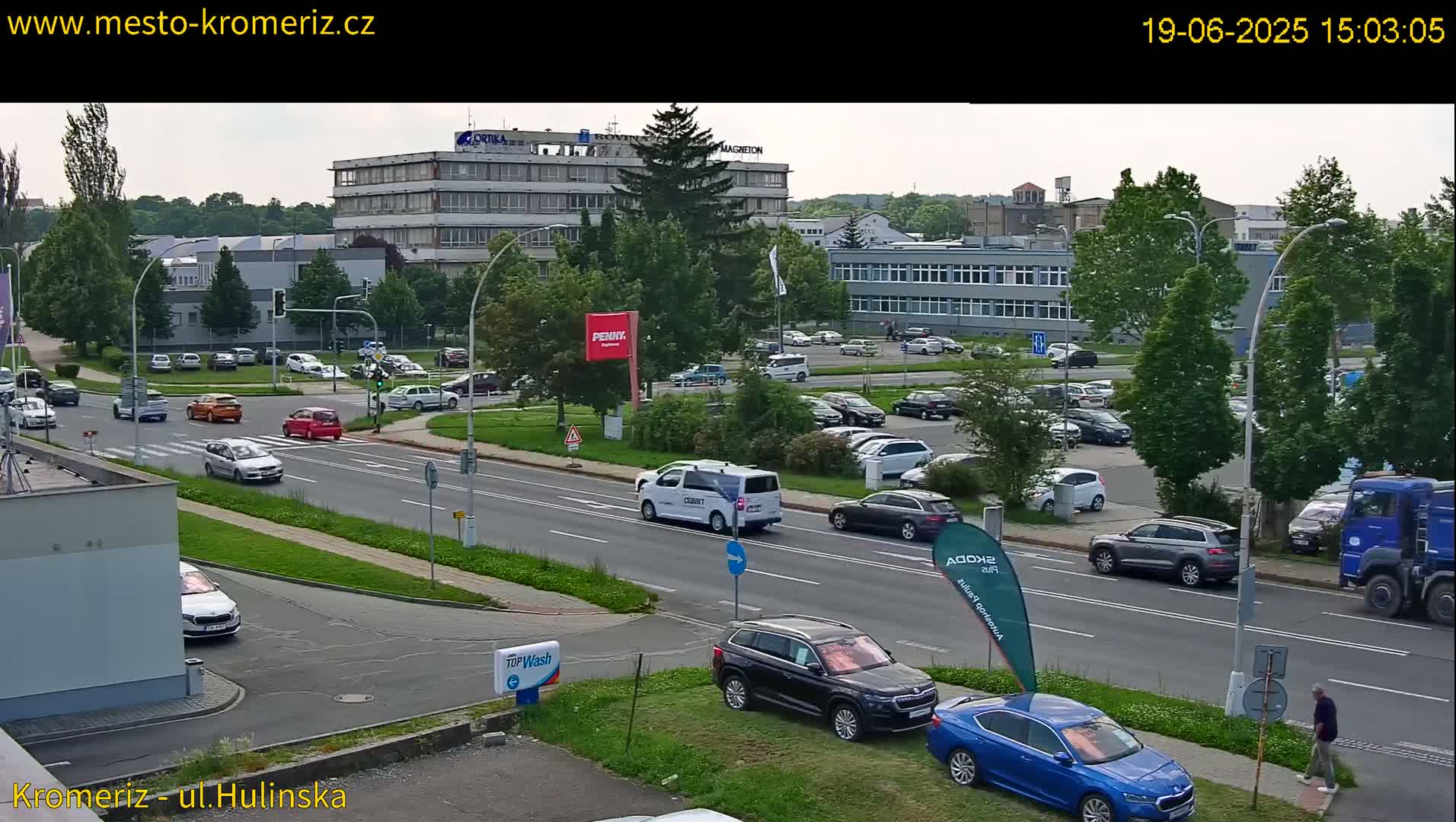 A partly sunny street scene shows several cars driving on a road lined with green landscaping, buildings, and a large parking lot filled with vehicles.