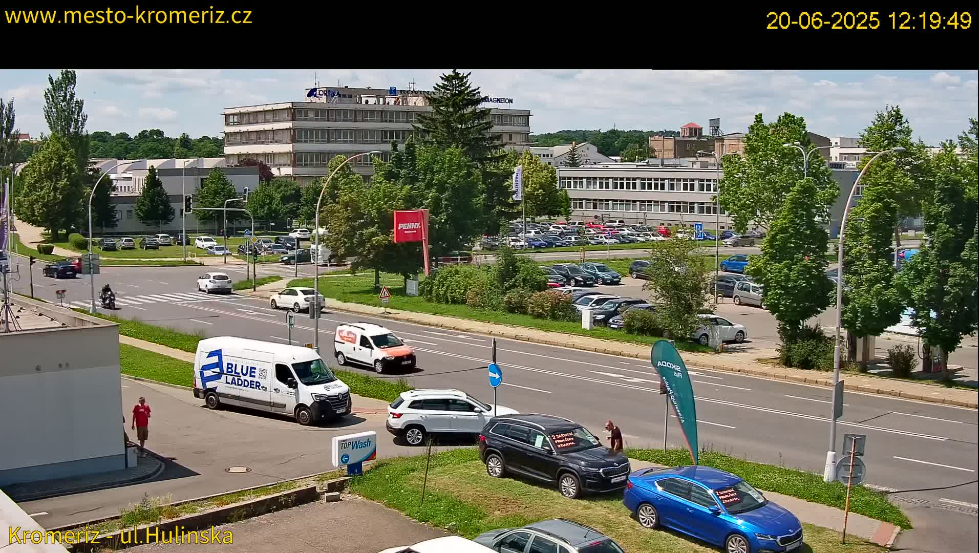 A sunny day reveals a street scene with several cars parked in a lot and driving on a road that passes by office buildings and some pedestrians.