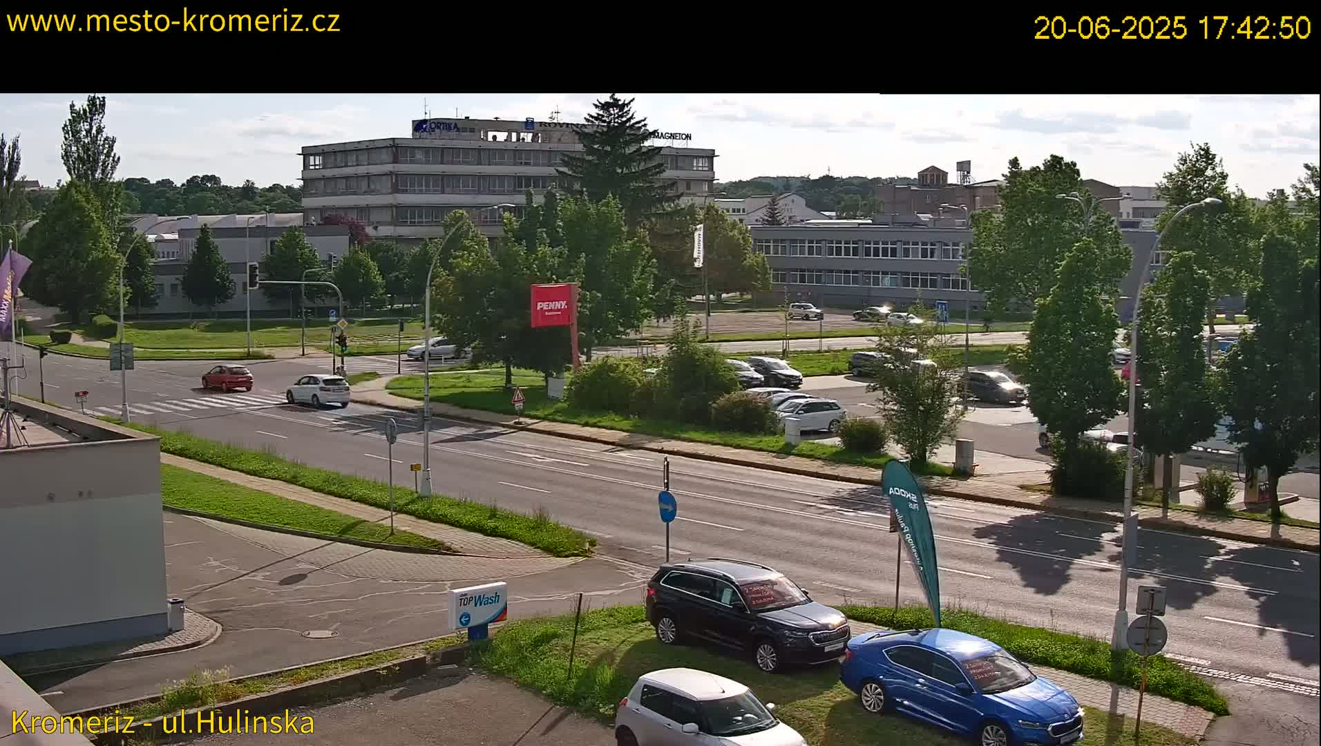 A sunny day view of a street scene with several cars parked and driving, along with buildings and green trees.