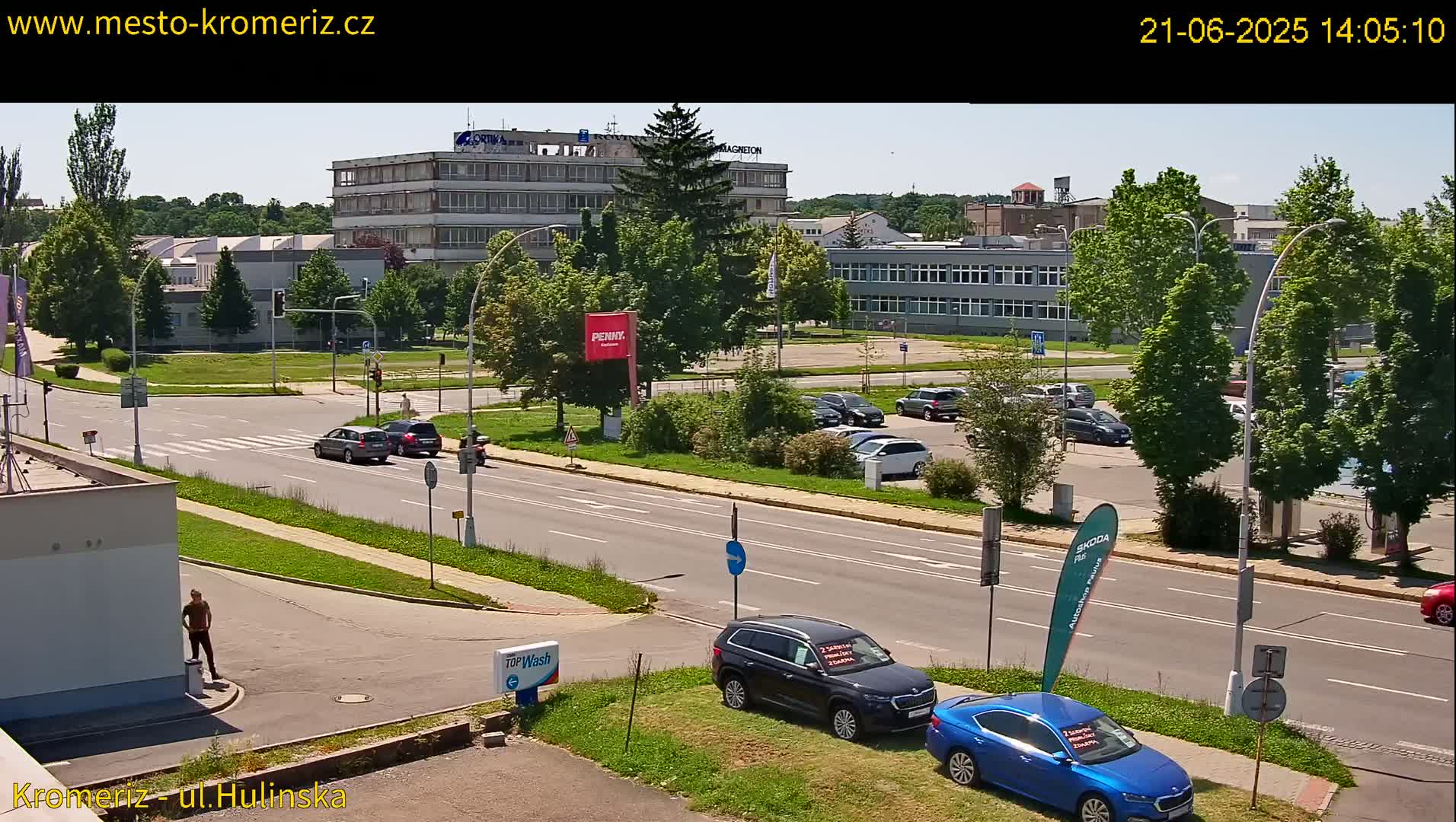 A sunny day view of a city street with several cars parked in a lot and a few driving on the road, near multi-story buildings with lush green trees.