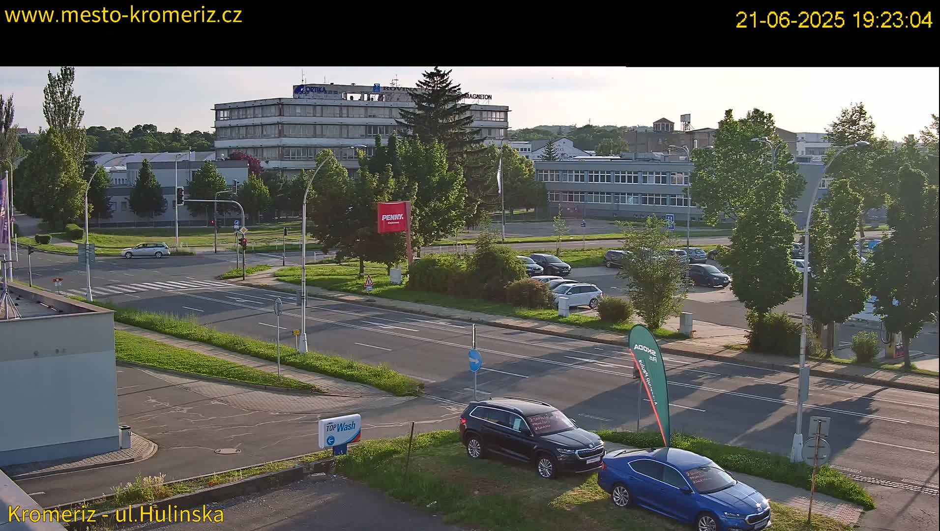 A street scene at dusk shows a multi-lane road with cars parked in a lot and driving on the road, several buildings in the background, and lush green trees lining the streets.