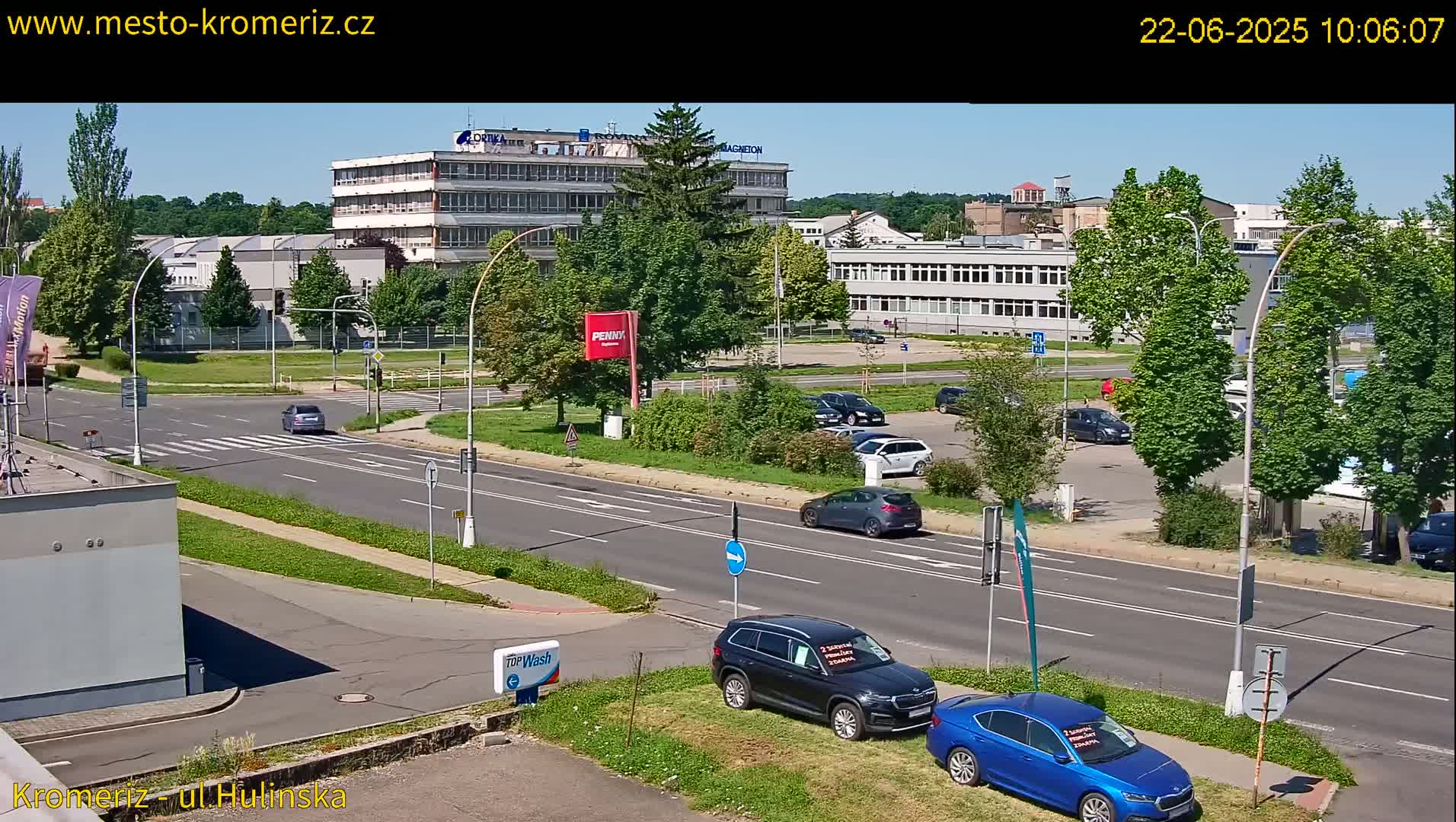 A sunny day reveals a street scene with several cars parked and driving near multi-story buildings and lush green trees.