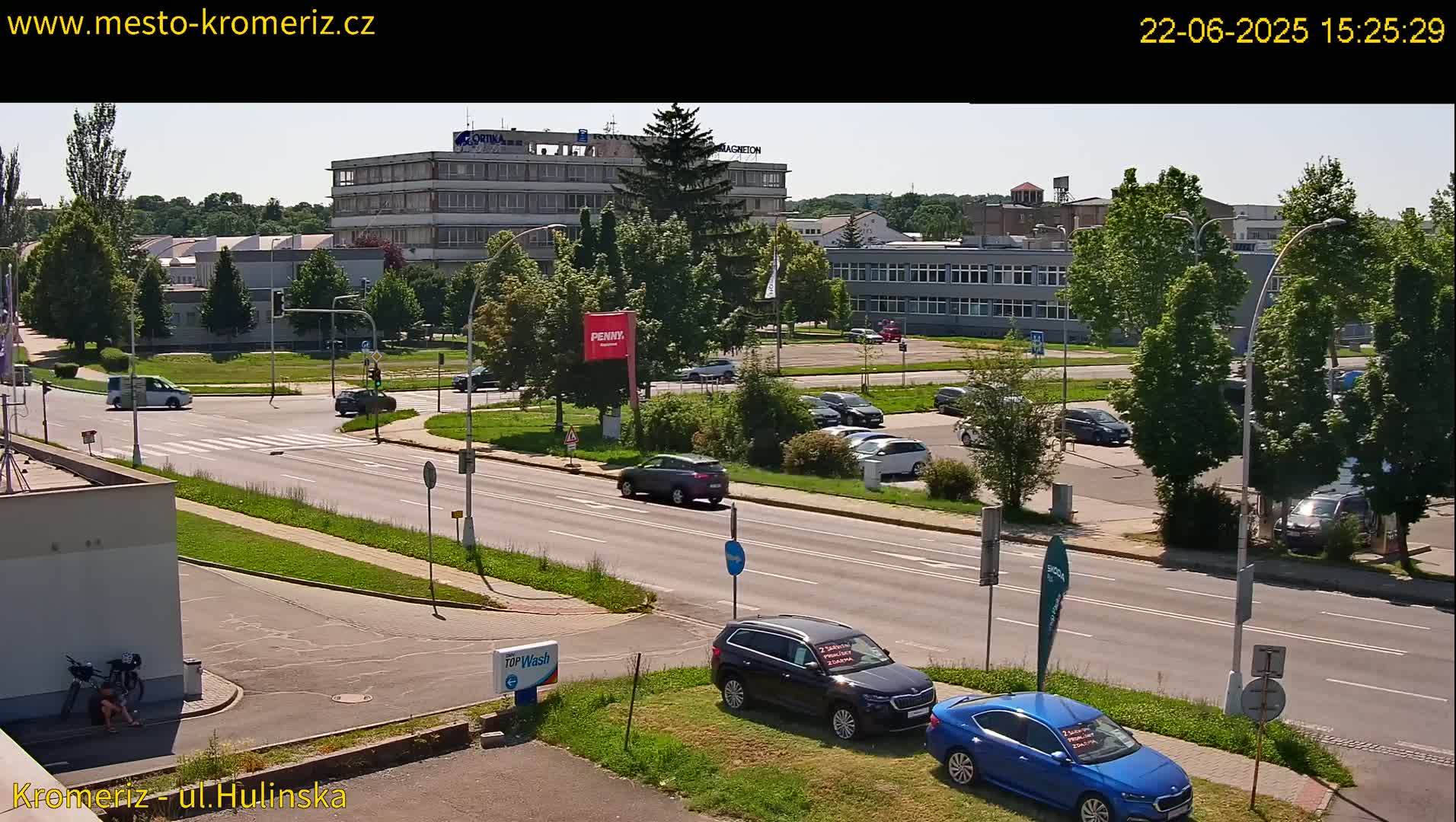 A sunny, daytime view of a street scene with several cars parked in a lot and others driving on the road, passing buildings and greenery.