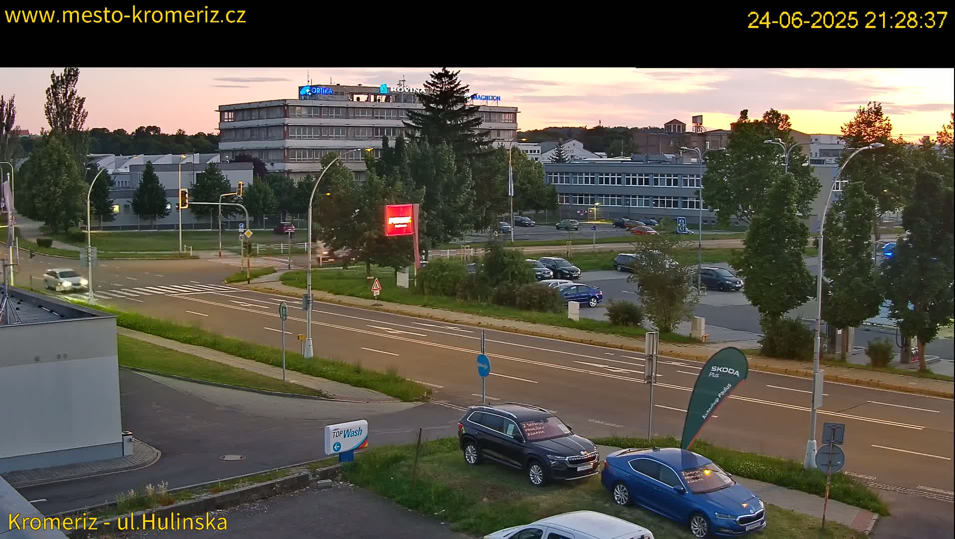 A twilight scene shows a multi-lane road with cars parked and driving, near a large industrial building and car dealership, under a partly cloudy sky.