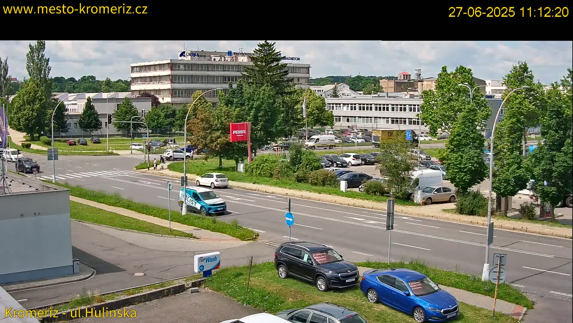 A sunny day shows a multi-lane road with several cars parked and driving, alongside buildings and lush green trees.
