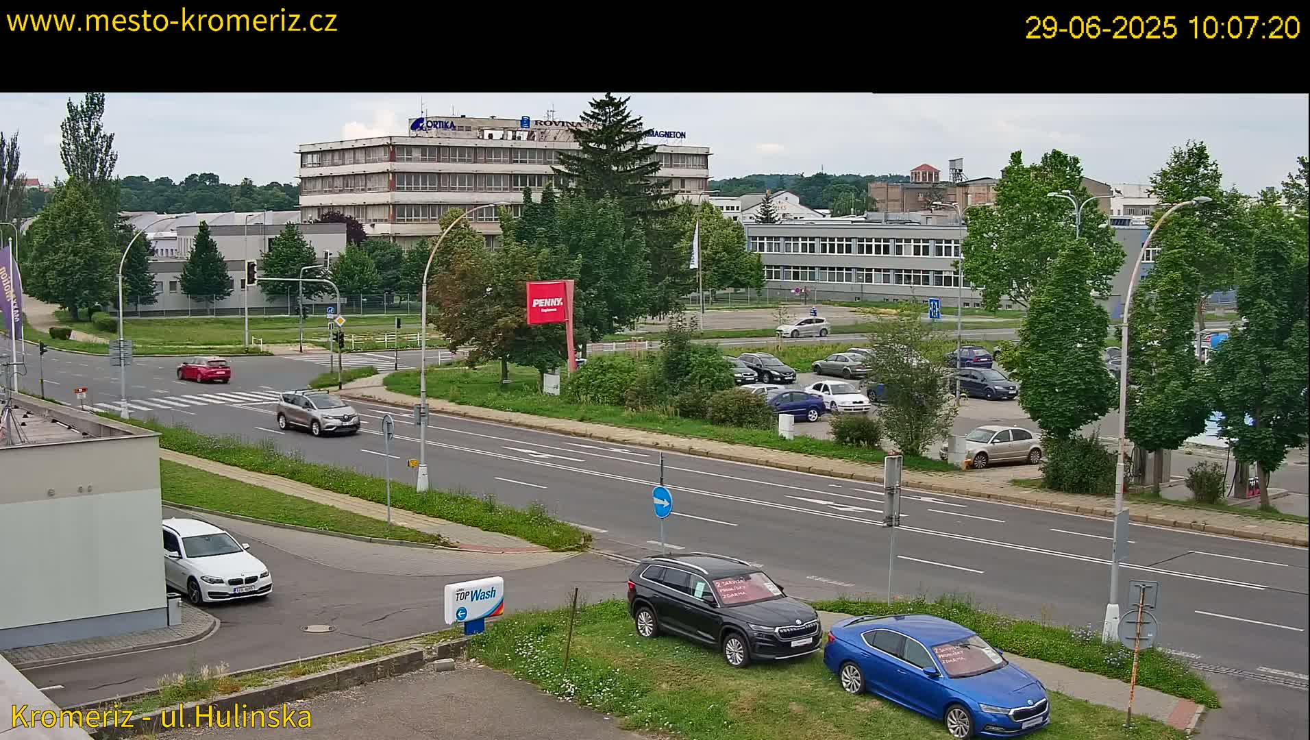 A mostly sunny day shows a street scene with several cars parked and driving near multi-story buildings and a car dealership.