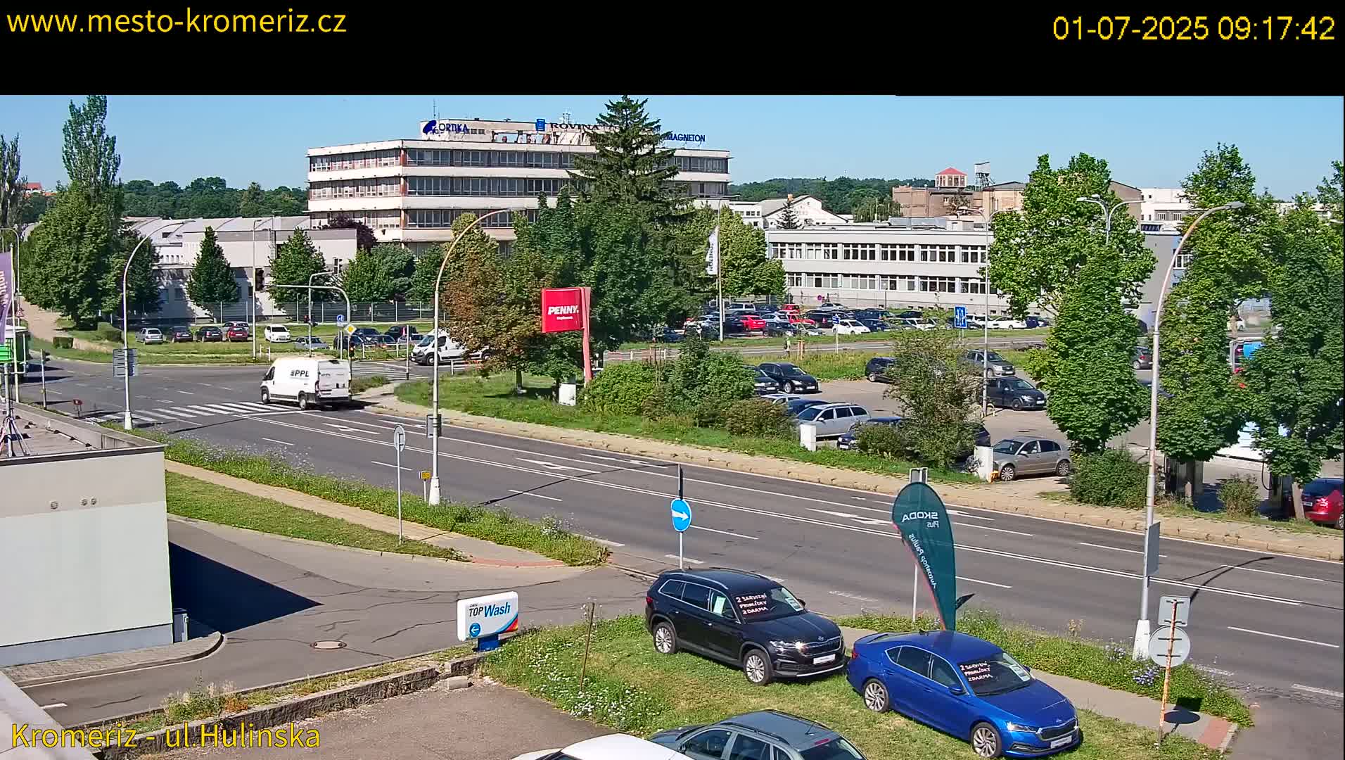 A sunny day reveals a view of a multi-story building complex and adjacent parking lot, with several cars parked, and a road with a white van driving by.