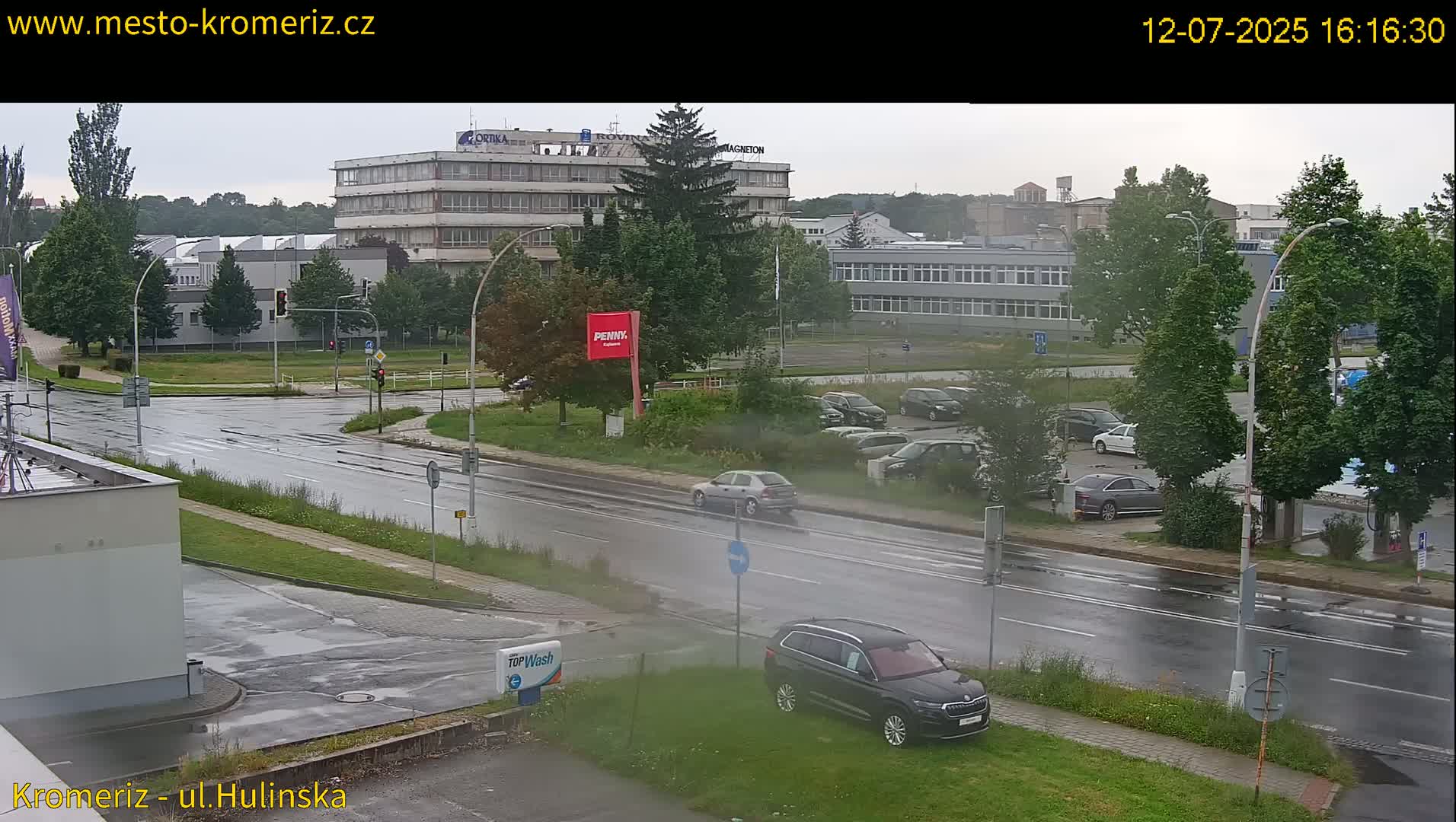 A sunny day reveals a view of a multi-story building complex and adjacent parking lot, with several cars parked, and a road with a white van driving by.