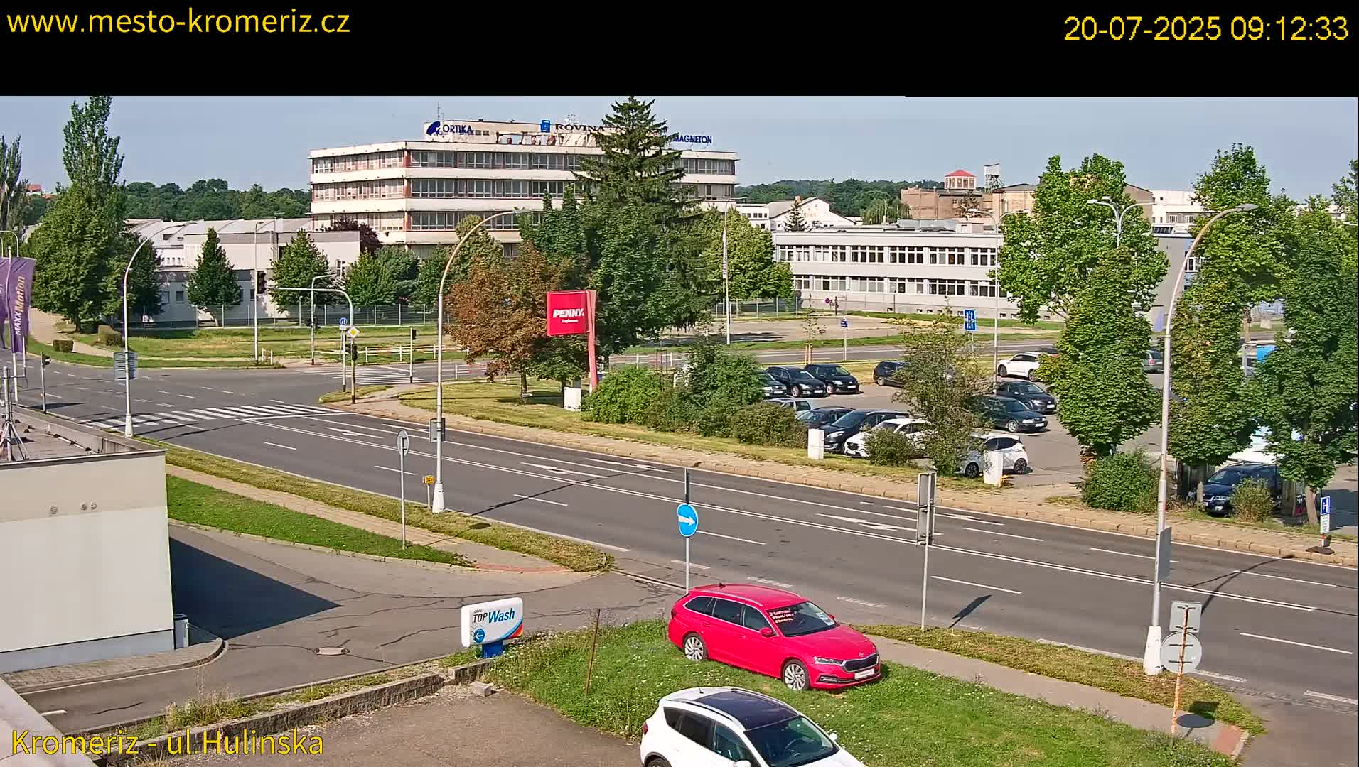 A sunny day reveals a wide road with cars parked in a lot across from several multi-story buildings and lush green trees.