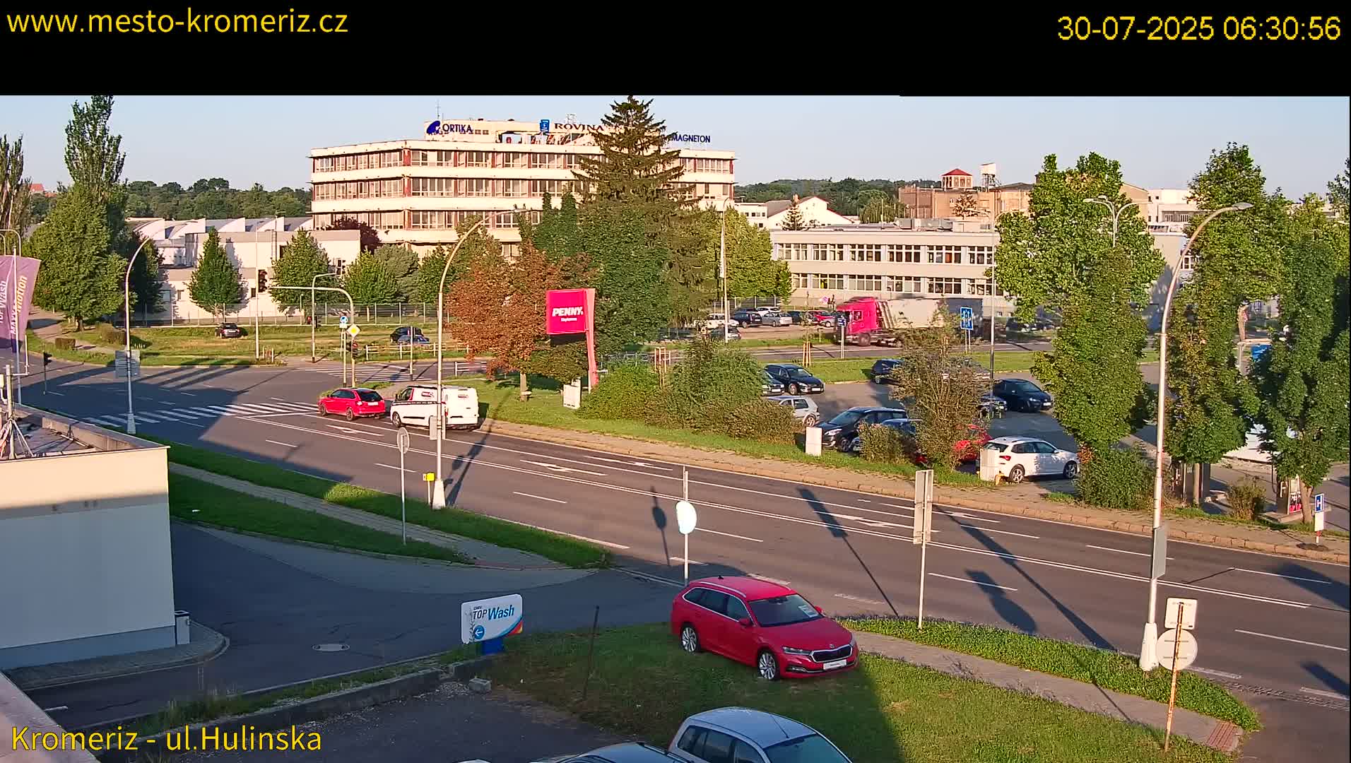 A sunny day reveals a wide road with cars parked in a lot across from several multi-story buildings and lush green trees.
