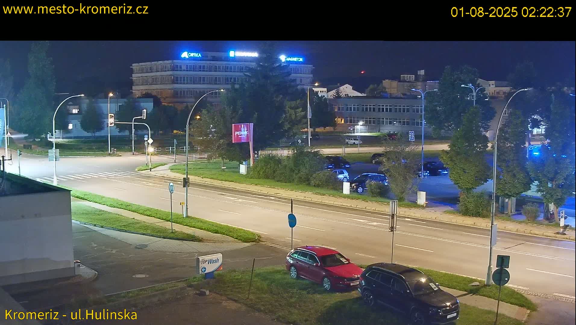 A nighttime street scene shows a multi-lane road with several cars parked and driving, passing in front of a large building complex under clear skies.