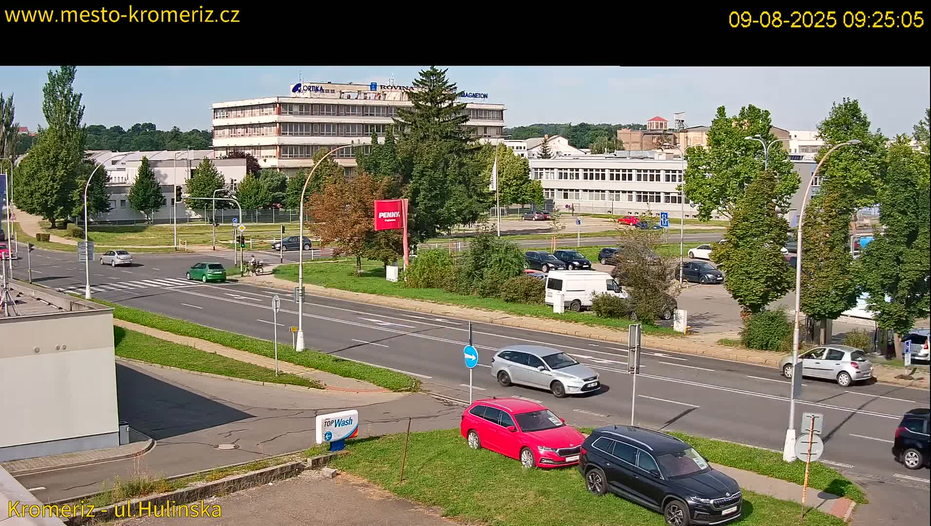 A sunny day shows a street intersection with several cars, a pedestrian crossing, and large buildings in the background.