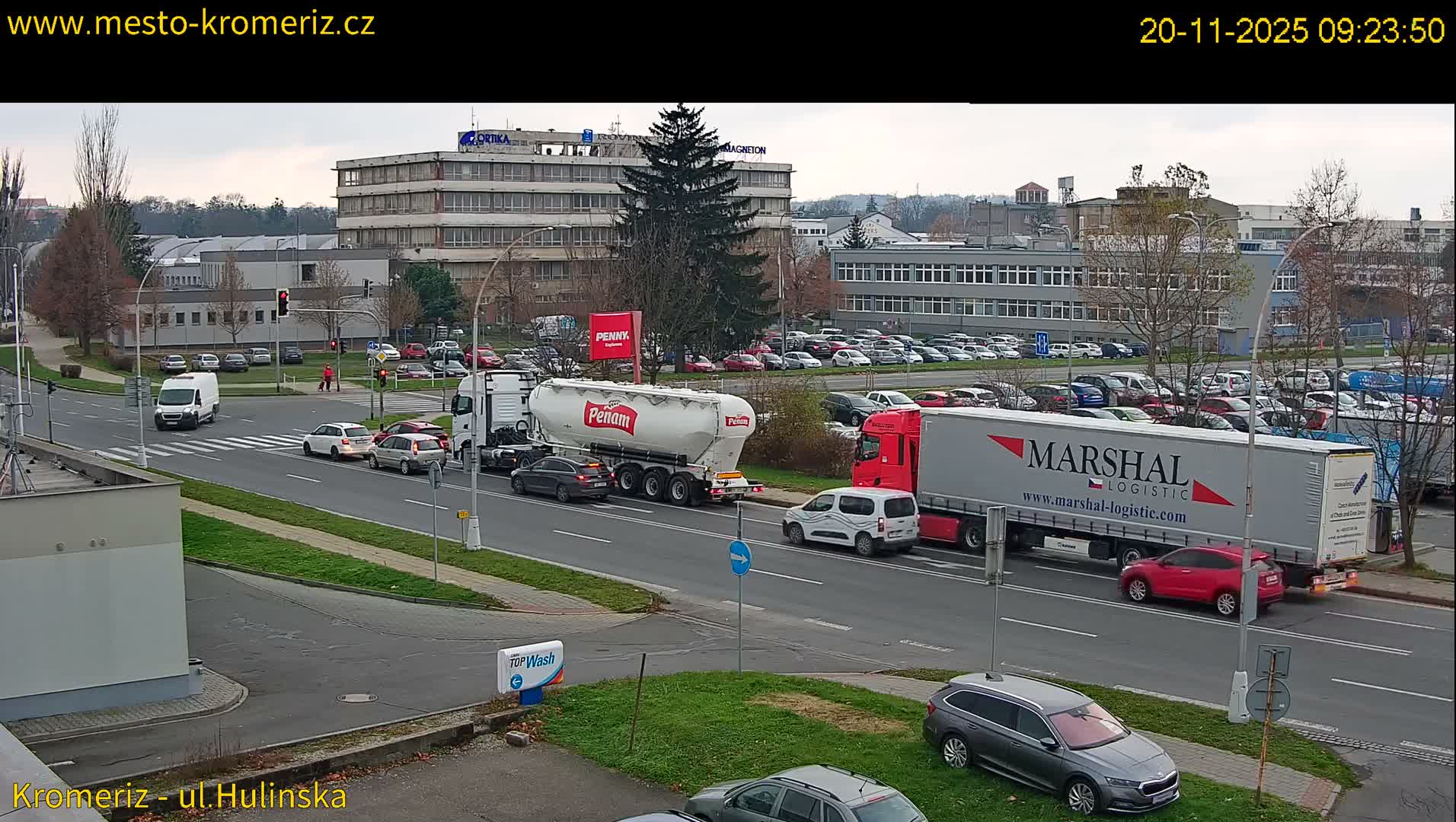 A wide-angle, elevated view captures a busy urban intersection with various vehicles including large trucks, cars, and a pedestrian, alongside multiple buildings and parking lots under a uniformly overcast sky.