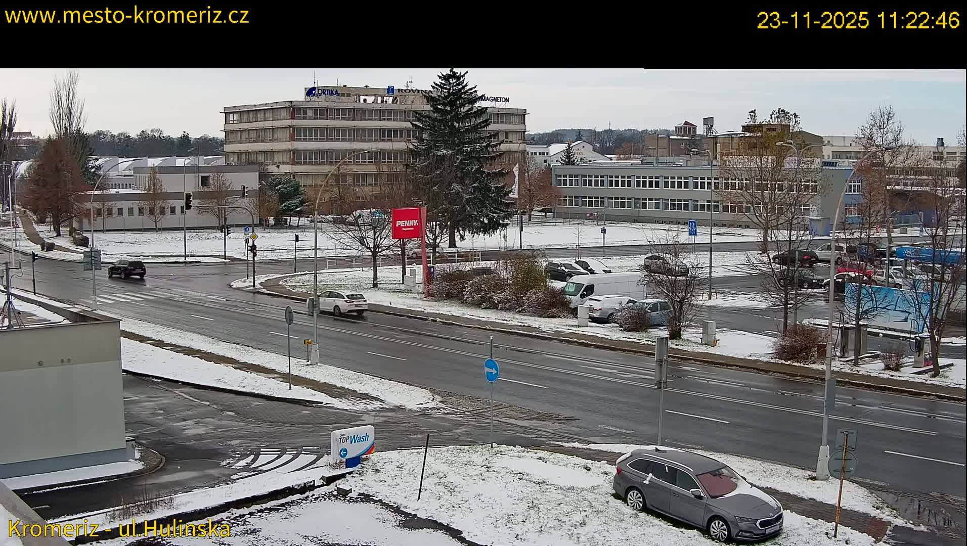 An elevated view captures an urban intersection and surrounding streets lightly covered in snow under an overcast sky, with several buildings, numerous bare trees, a prominent evergreen, and cars moving or parked along the snowy roadsides.