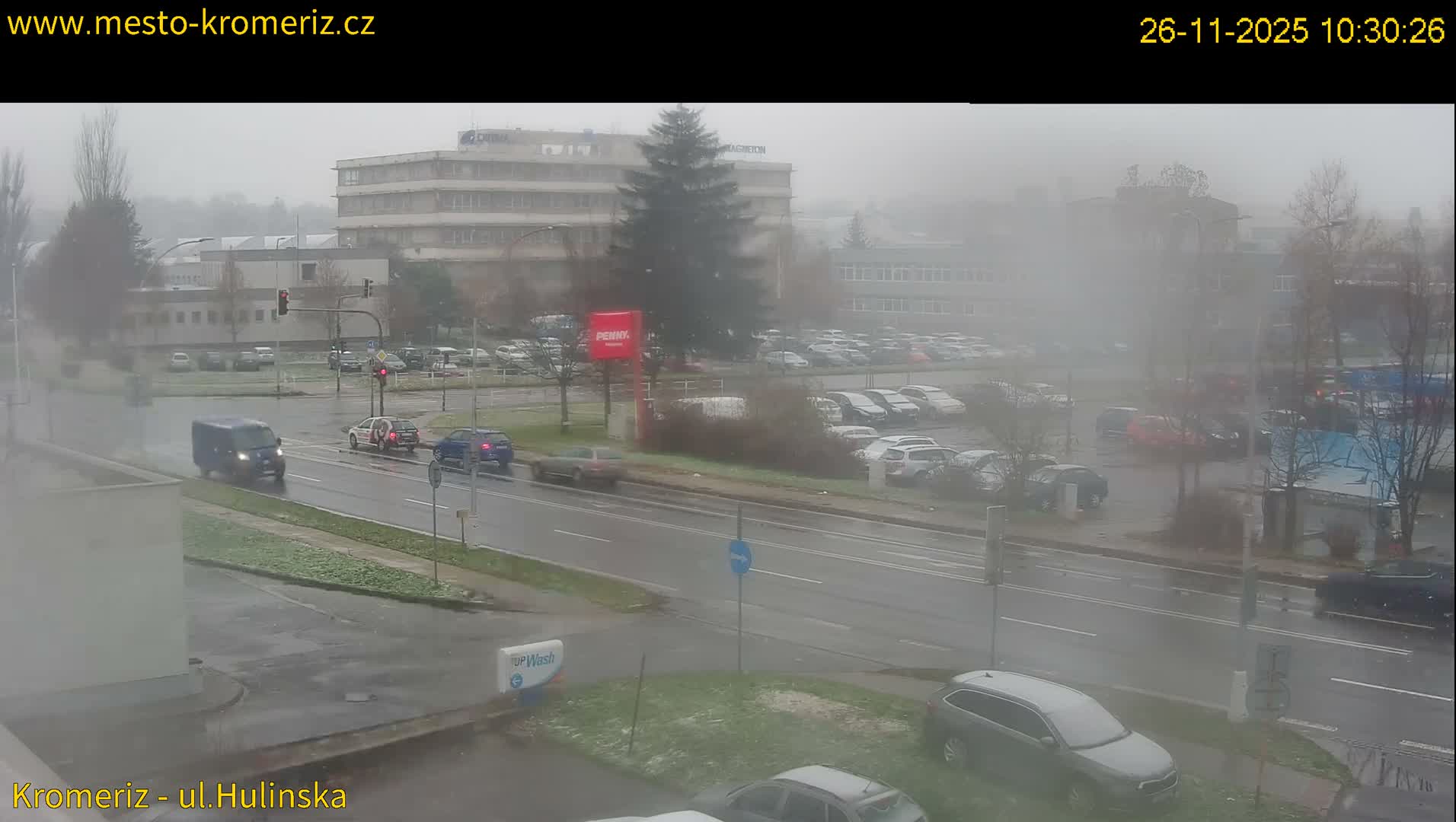 A misty and overcast urban street scene displays wet roads with cars driving and parked, surrounded by buildings and bare trees, with scattered patches of frost or snow on the ground.