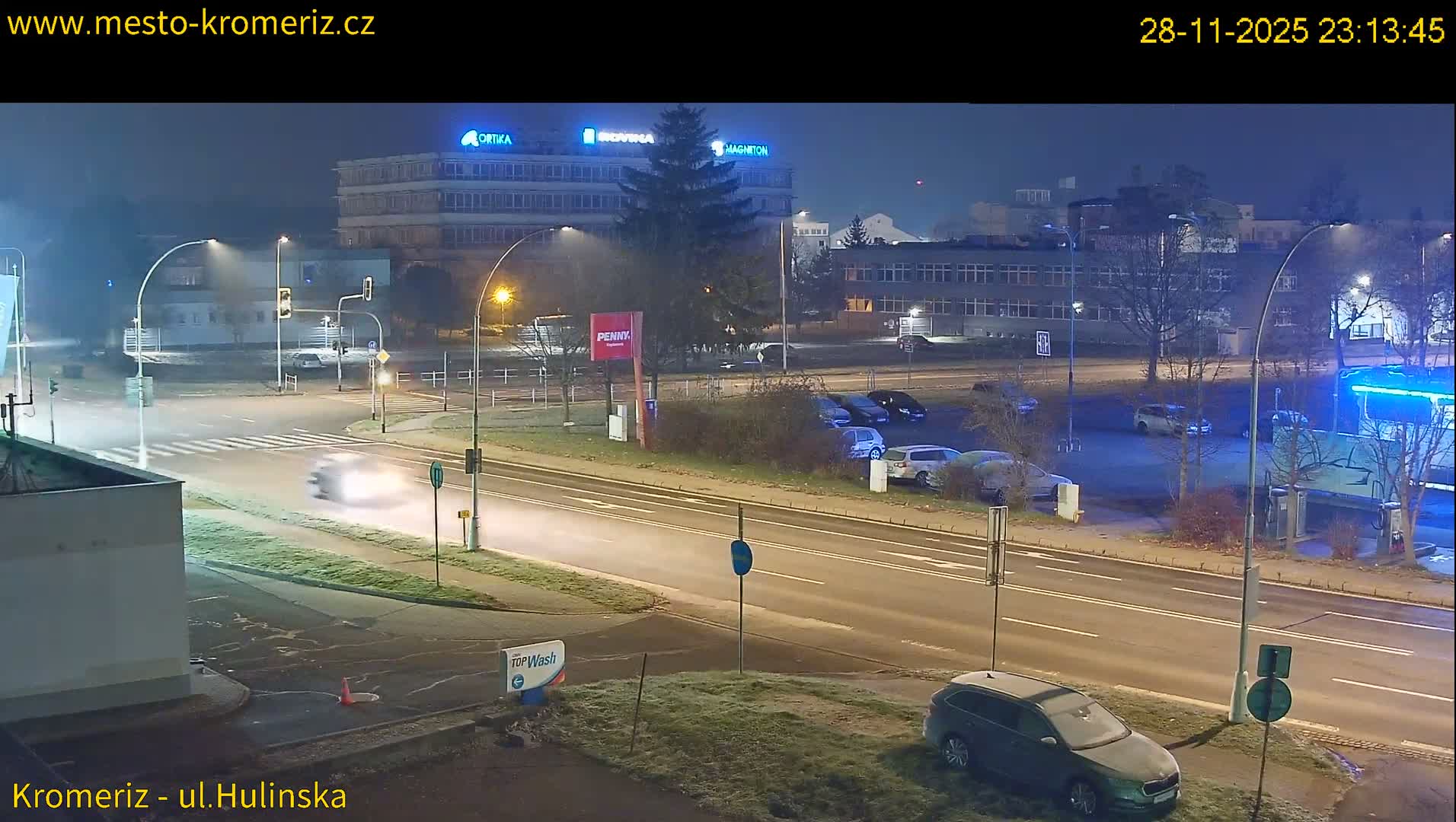 The image displays a clear, cold urban intersection at night, with frost visible on the ground and parked vehicles, illuminated by streetlights and featuring a blurred car in motion with various buildings in the background.