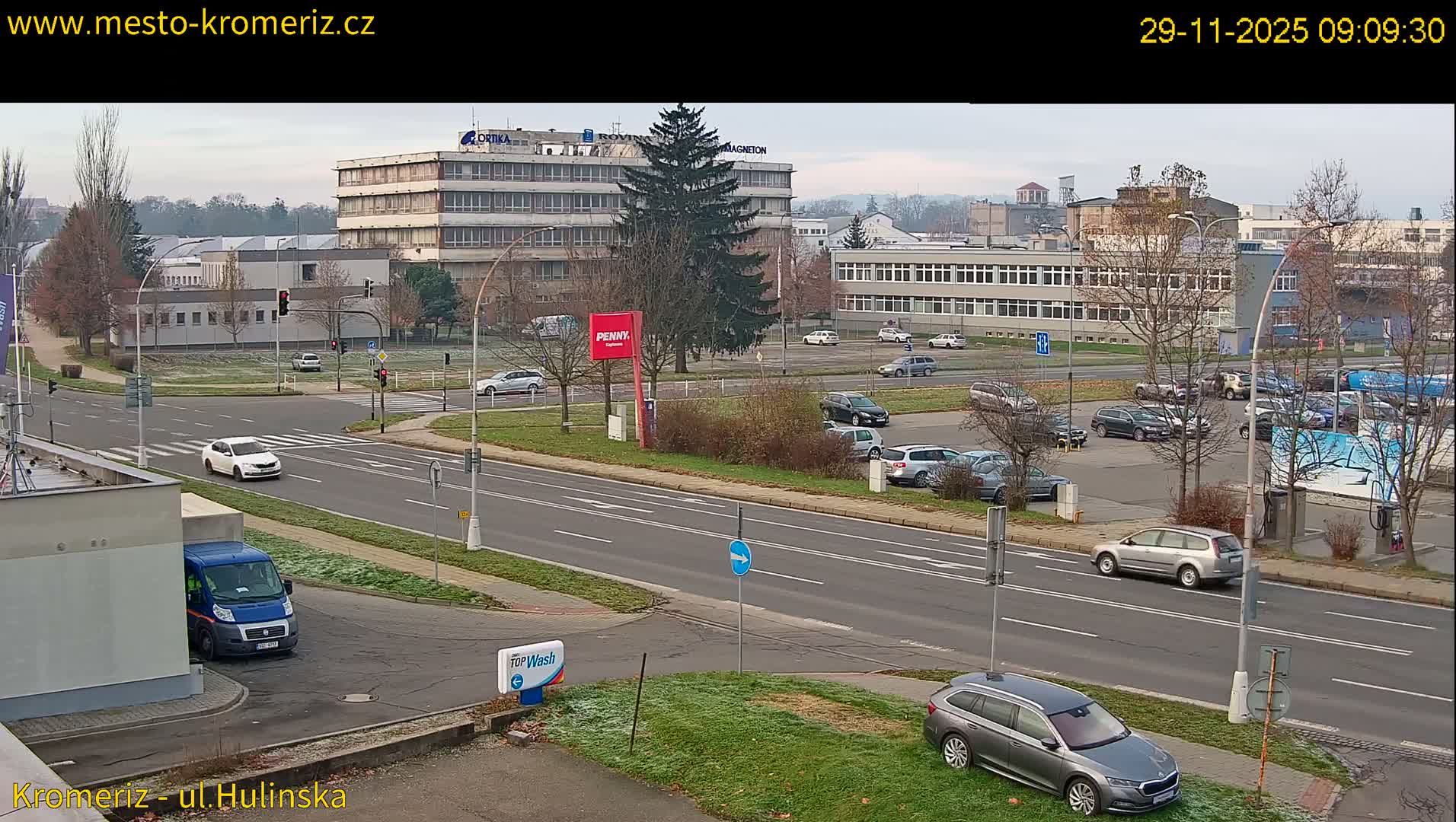 A wide-angle view captures an urban intersection with multiple cars driving and parked along roads bordered by frosted grass verges, bare trees, and various buildings under an overcast sky.