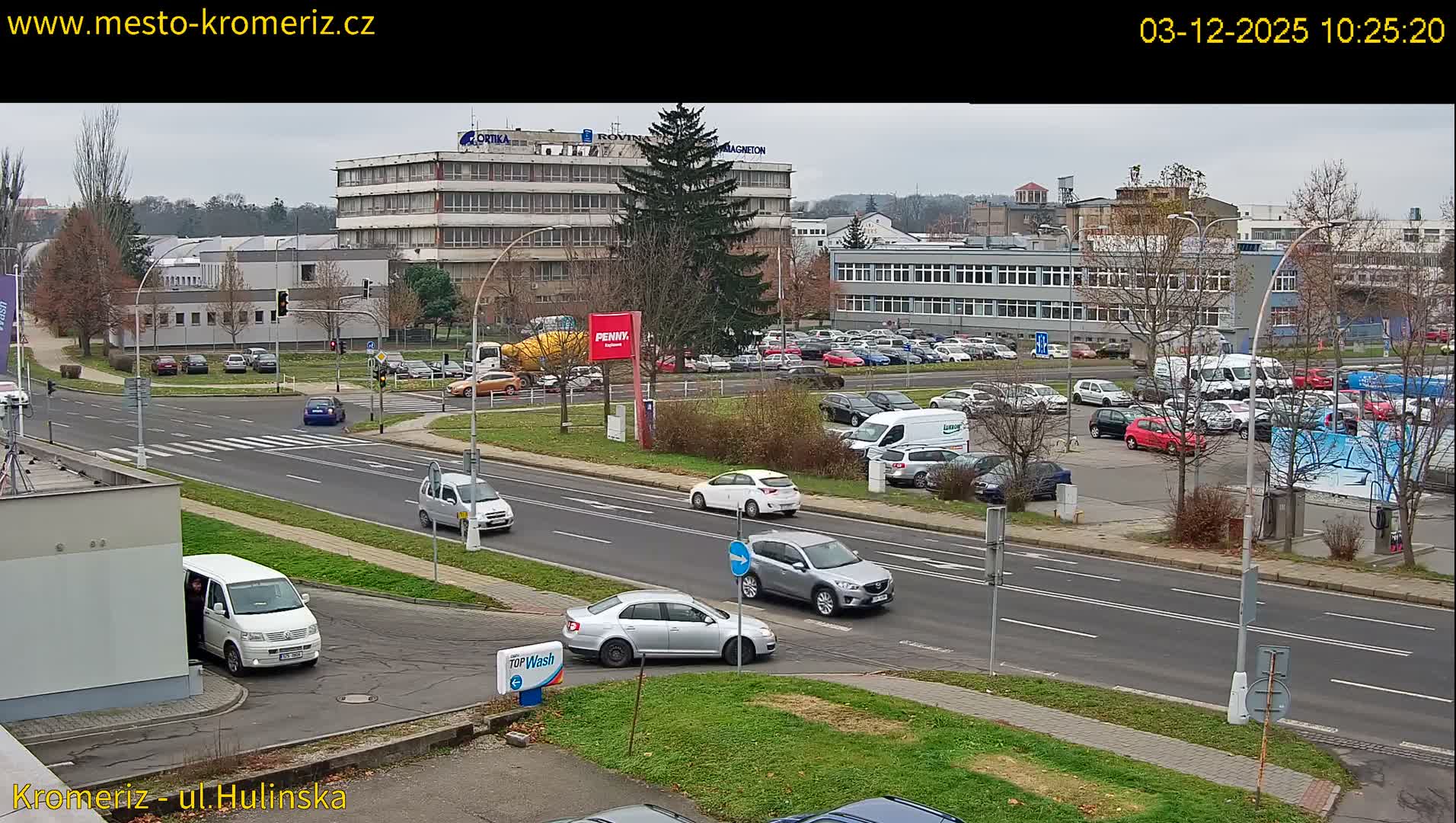 An overcast daytime scene shows multiple cars driving and parked along a multi-lane road at an urban intersection, surrounded by various buildings and mostly bare deciduous trees.