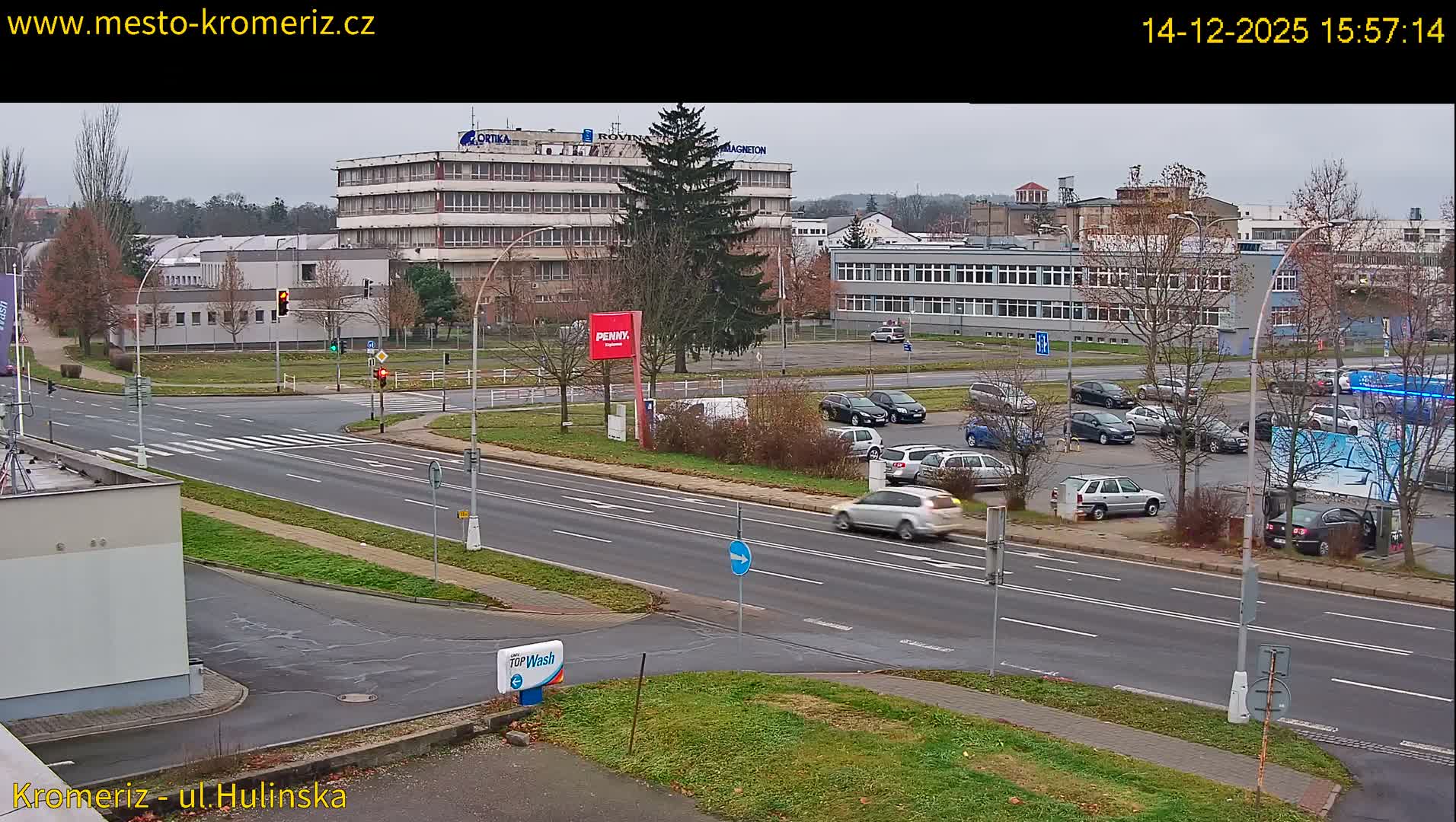 An overcast daytime scene shows multiple cars driving and parked along a multi-lane road at an urban intersection, surrounded by various buildings and mostly bare deciduous trees.