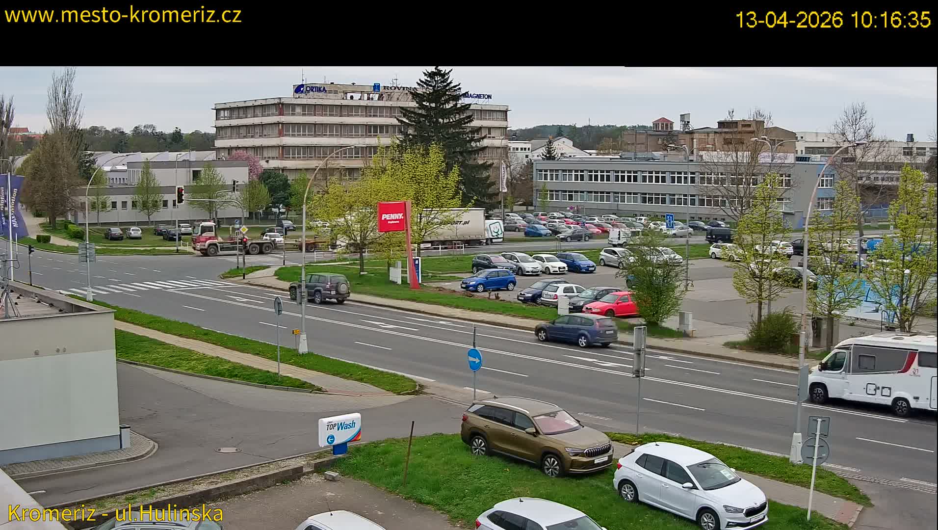 An overcast daytime scene shows multiple cars driving and parked along a multi-lane road at an urban intersection, surrounded by various buildings and mostly bare deciduous trees.