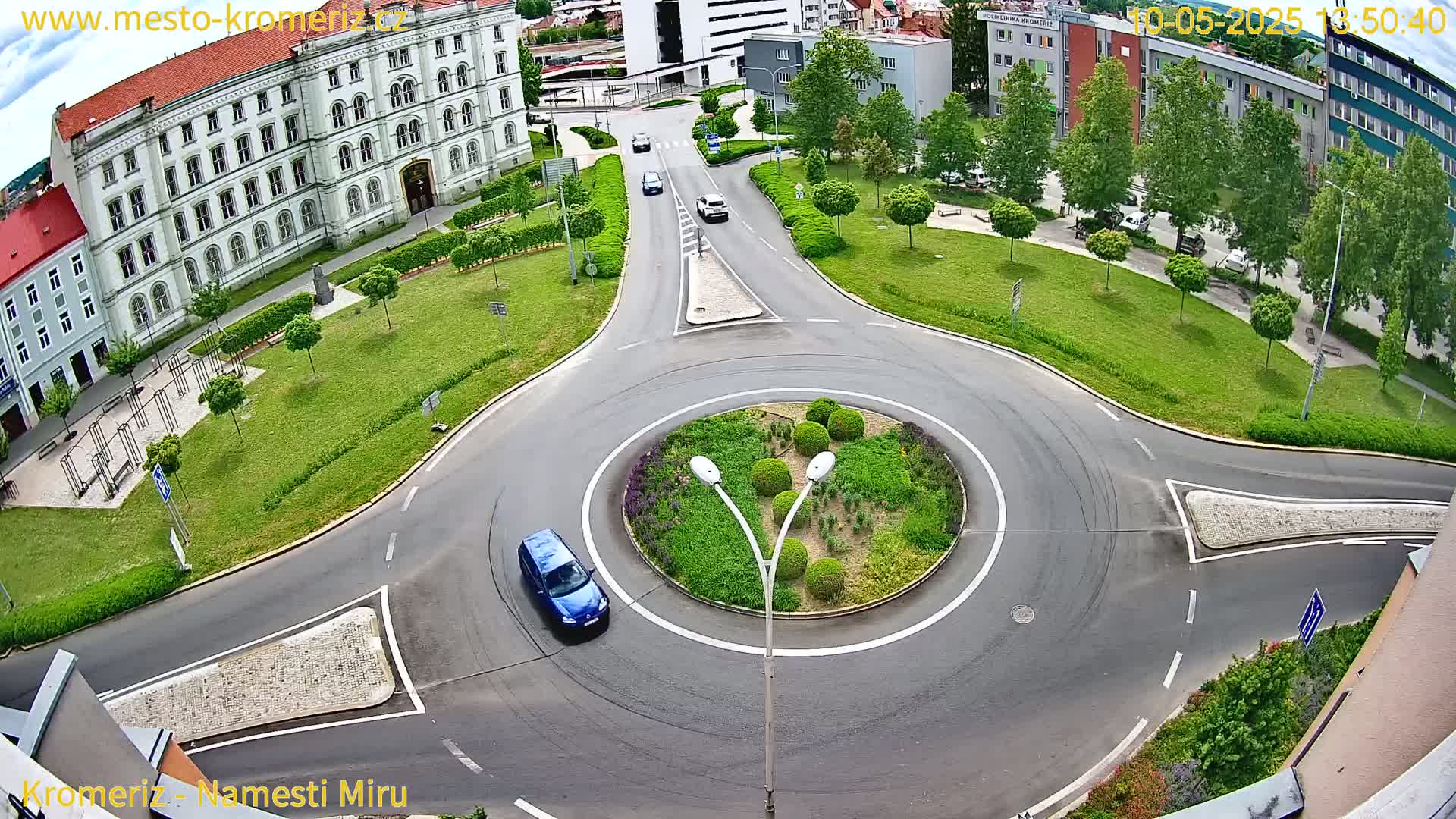 An aerial view of a roundabout in a city with a large building and green spaces, on a sunny day.