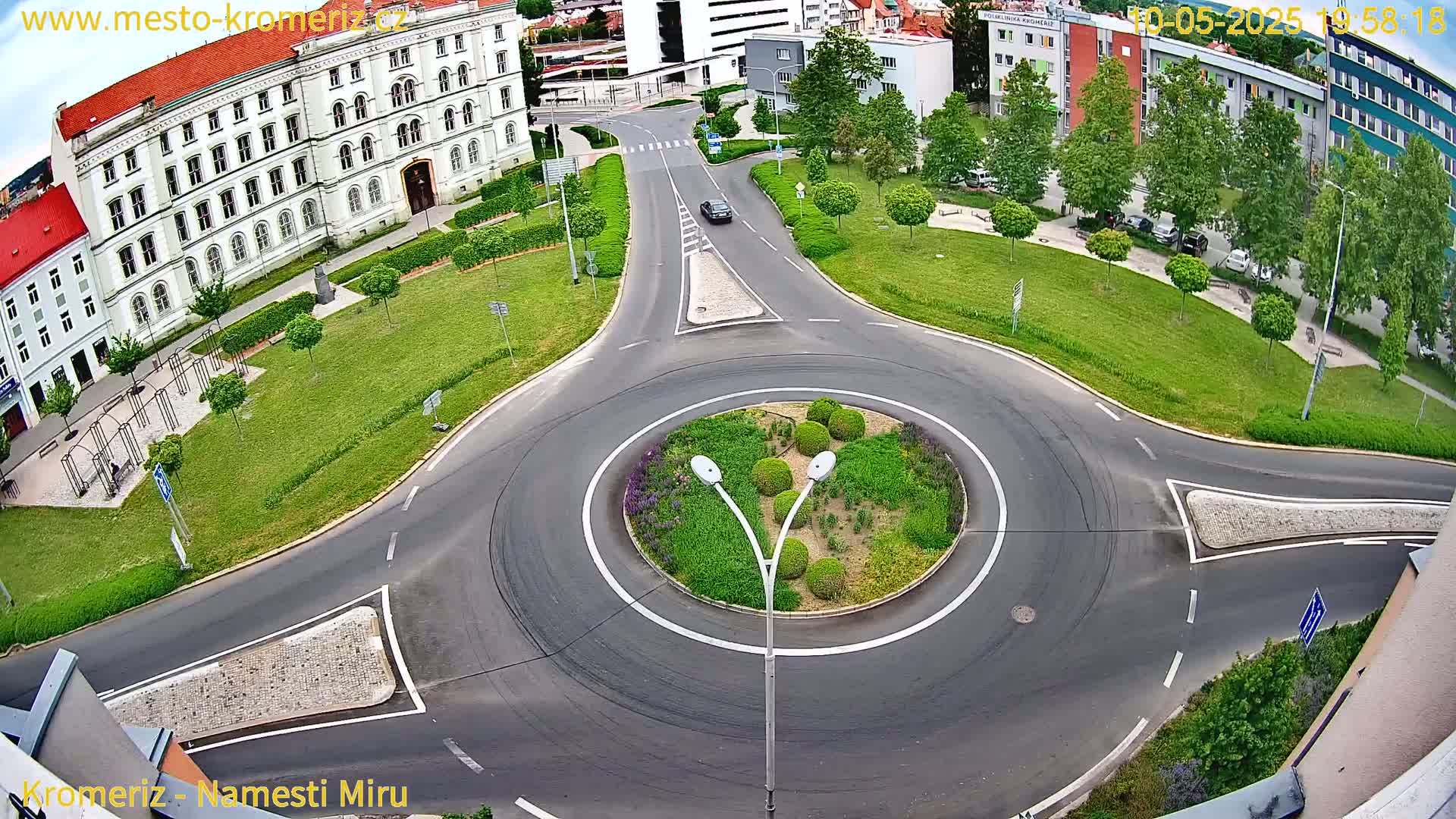 A car drives through a roundabout surrounded by green lawns, trees, and buildings on a sunny day.