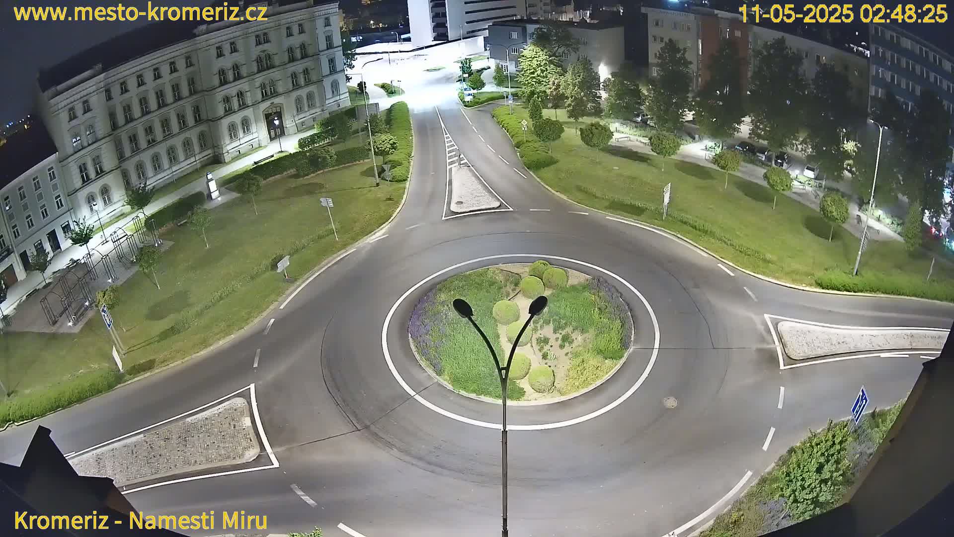 A nighttime overhead view of a roundabout surrounded by a grassy area with buildings and trees in the background.
