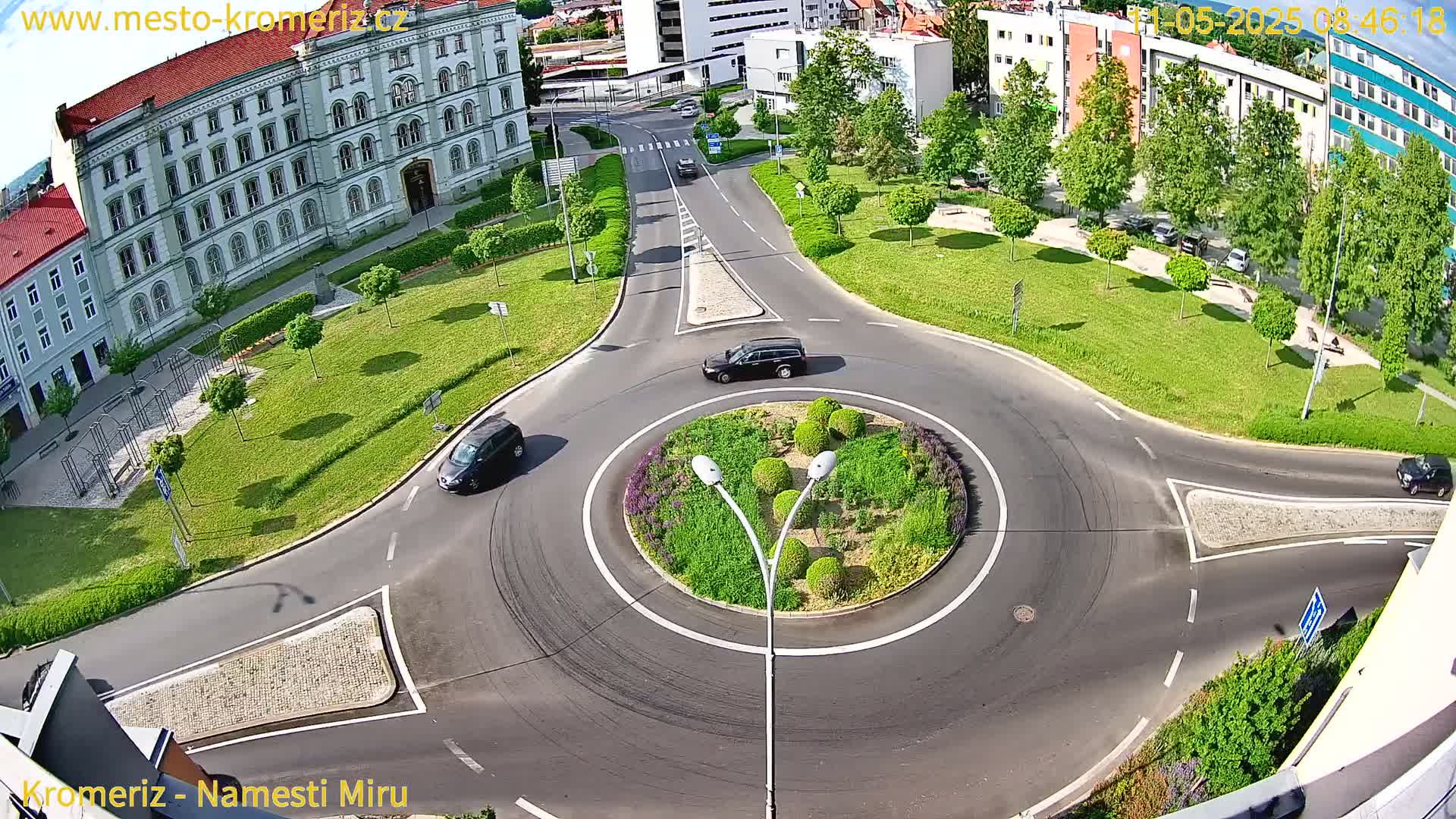 A sunny day shows a roundabout with several cars driving around it, situated in a town square with green grass and a large building in the background.