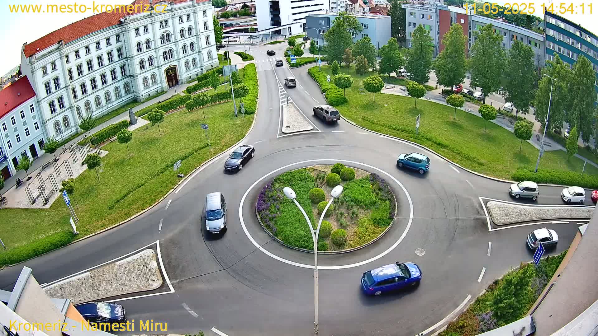 An aerial view shows a roundabout surrounded by green lawns, buildings, and several cars driving in a sunny day.