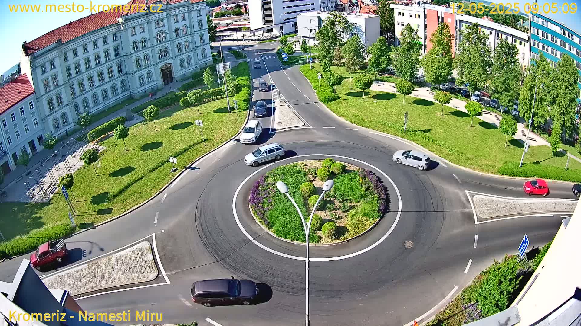 An aerial view shows a sunny day over a roundabout with several cars driving around a landscaped center island, near a large building and green park.