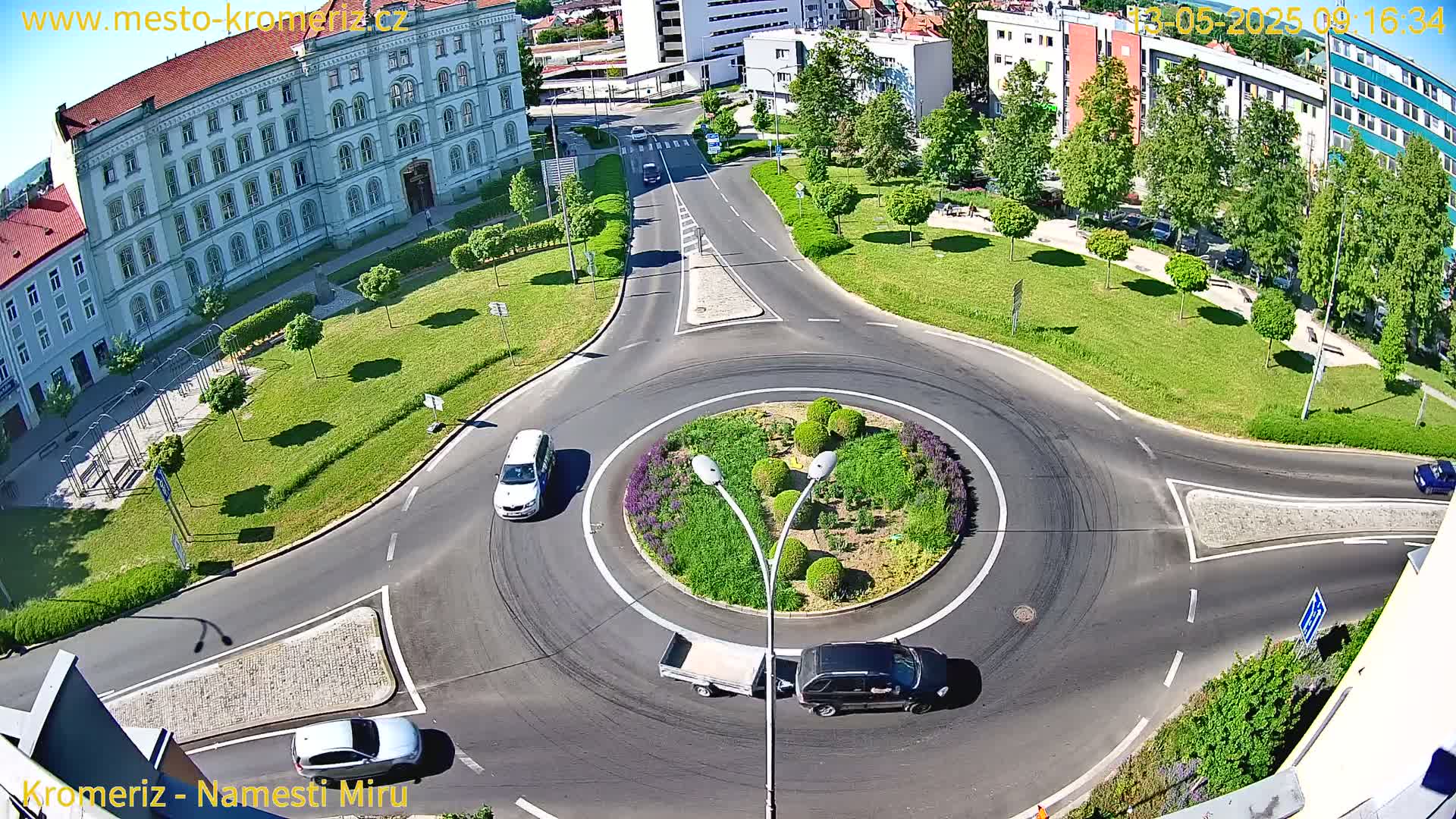 A sunny aerial view shows a roundabout with several cars, surrounded by a grassy park and buildings.
