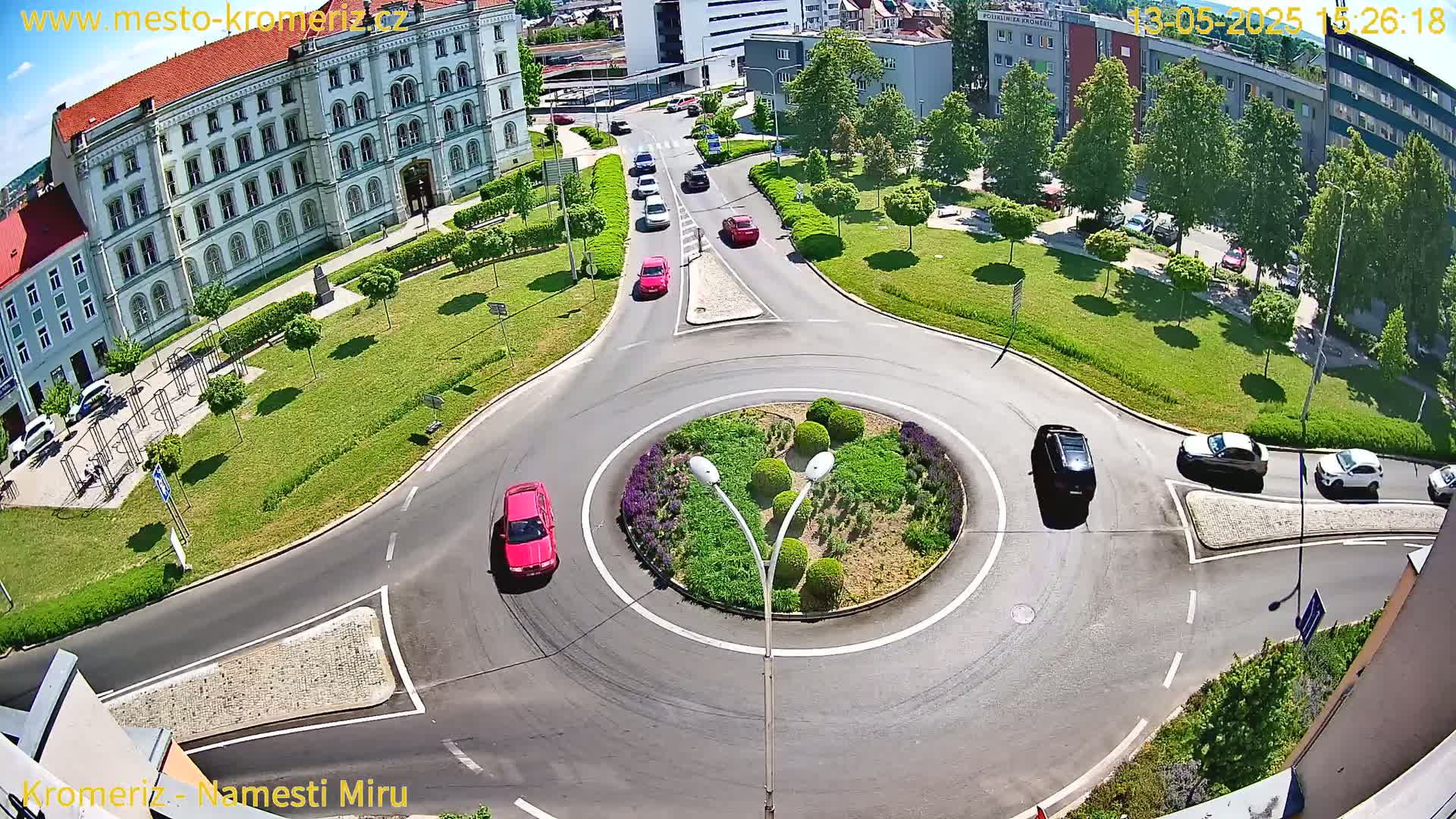 An aerial view shows a roundabout with several cars driving around it, next to a large building and a grassy area on a sunny day.