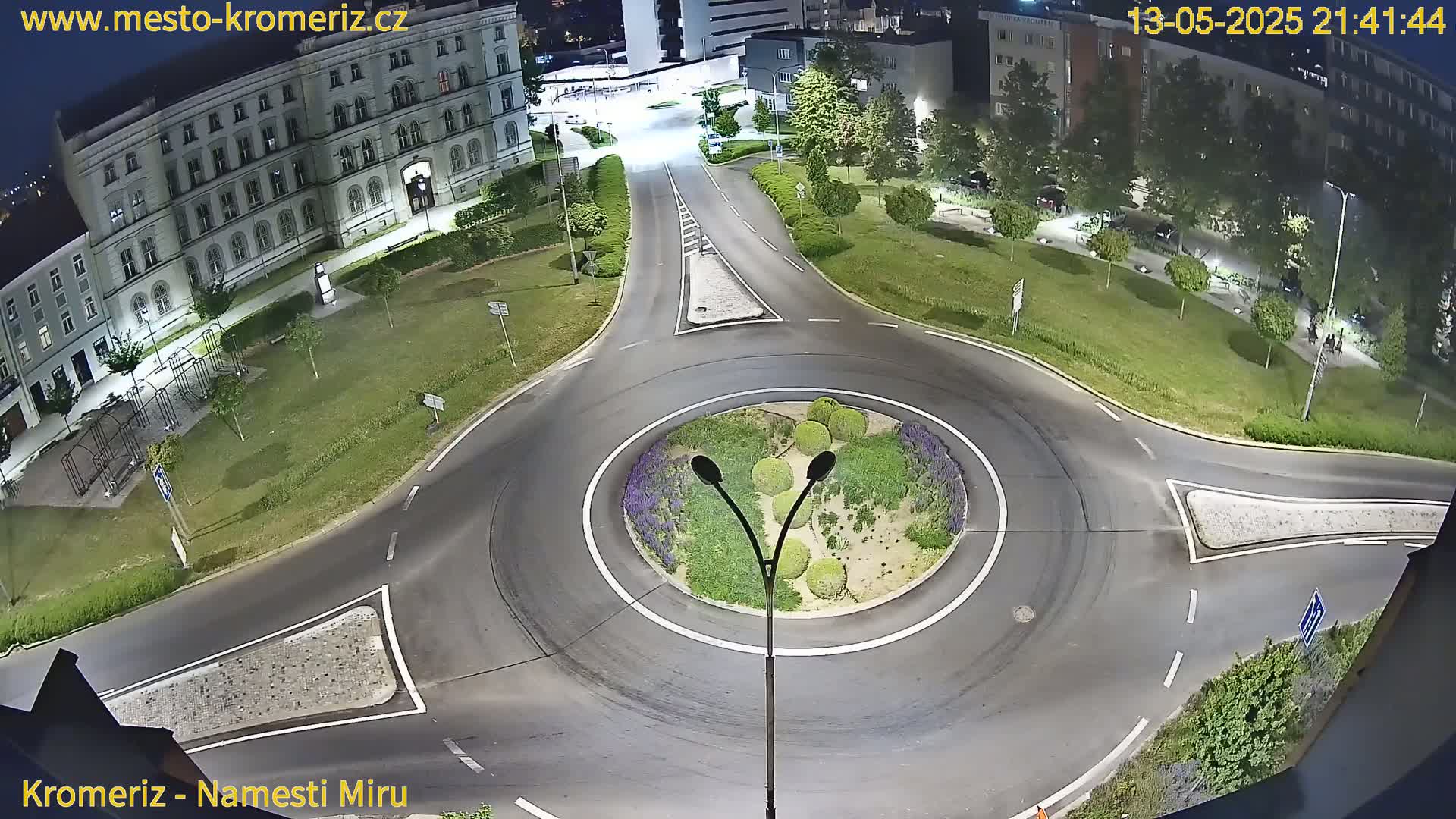 A nighttime aerial view shows a roundabout surrounded by a landscaped area and buildings, with a few cars visible on the roads.