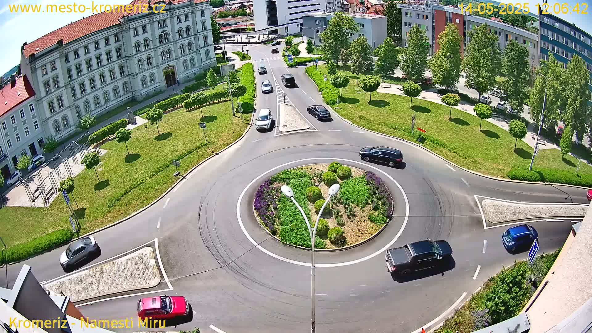 An aerial view shows a sunny day over a roundabout surrounded by green lawns and buildings, with several cars driving around and through it.