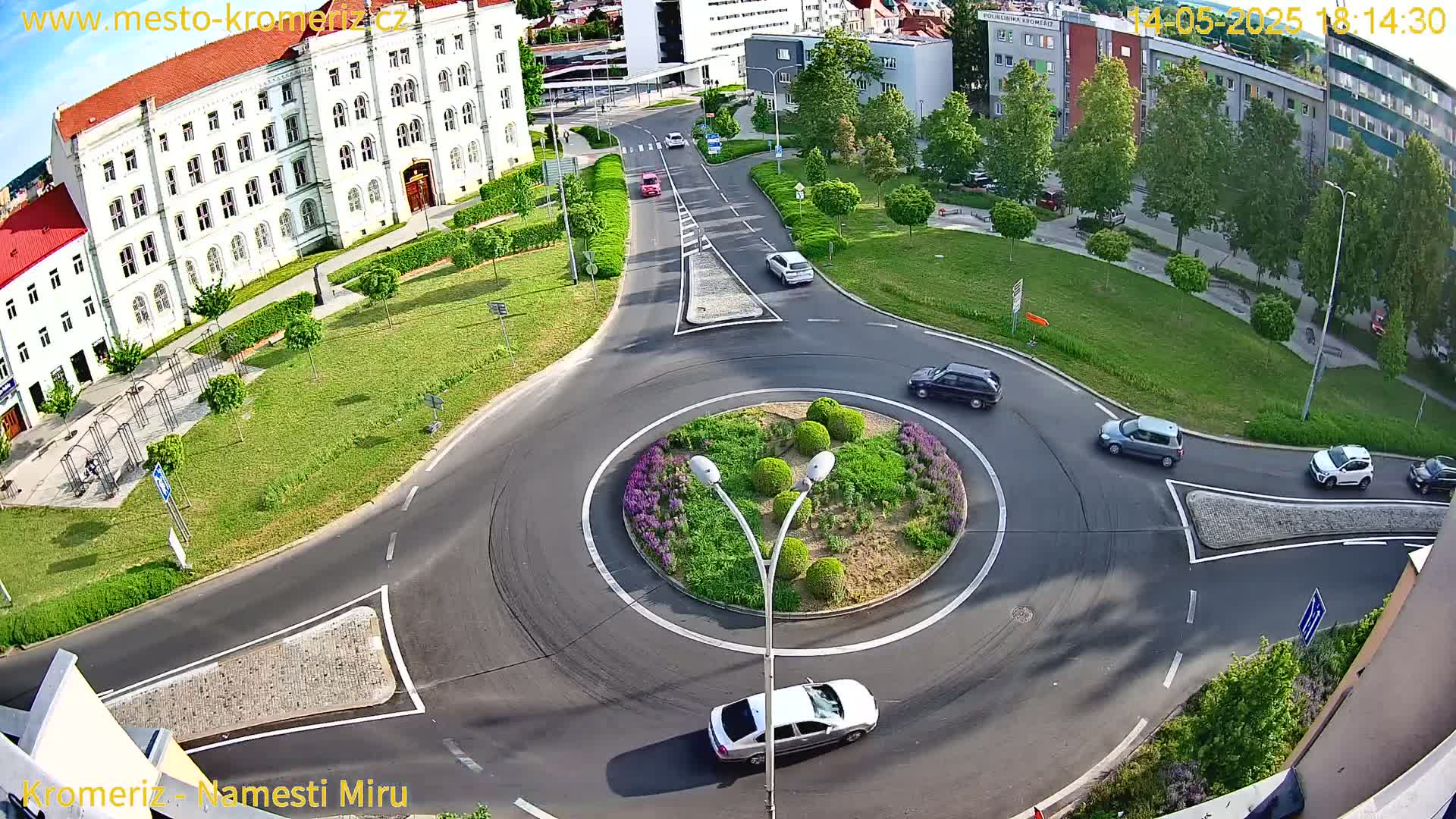 An aerial view of a traffic circle surrounded by green lawns and buildings, on a sunny day, with several cars circulating the roundabout.